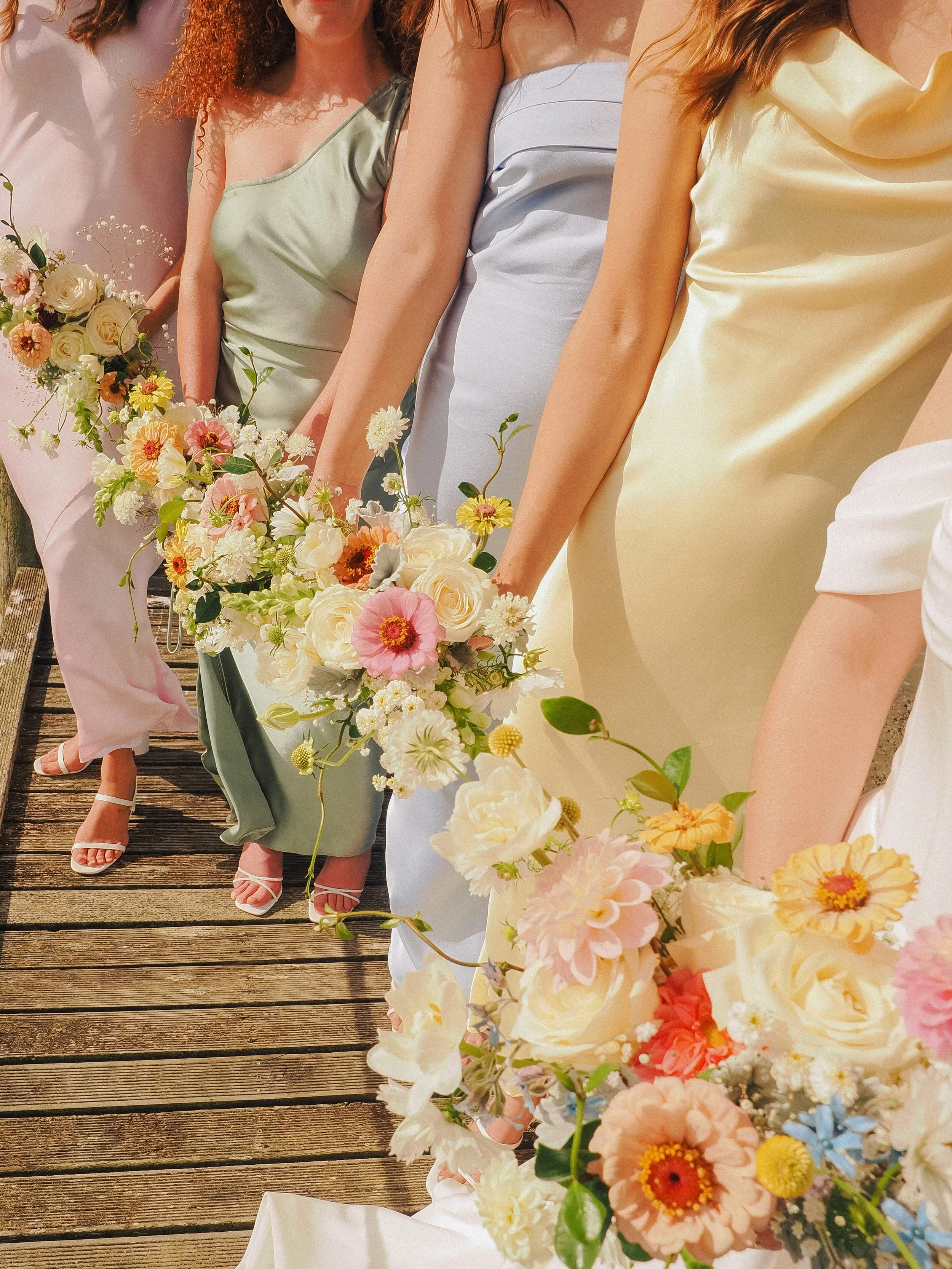 Group of women dressed in pastel-colored dresses holding bouquets of flowers on a wooden walkway.  Wedding Photography Waikato New Zealand. Wedding Photography Hamilton New Zealand. Northland Wedding Photography