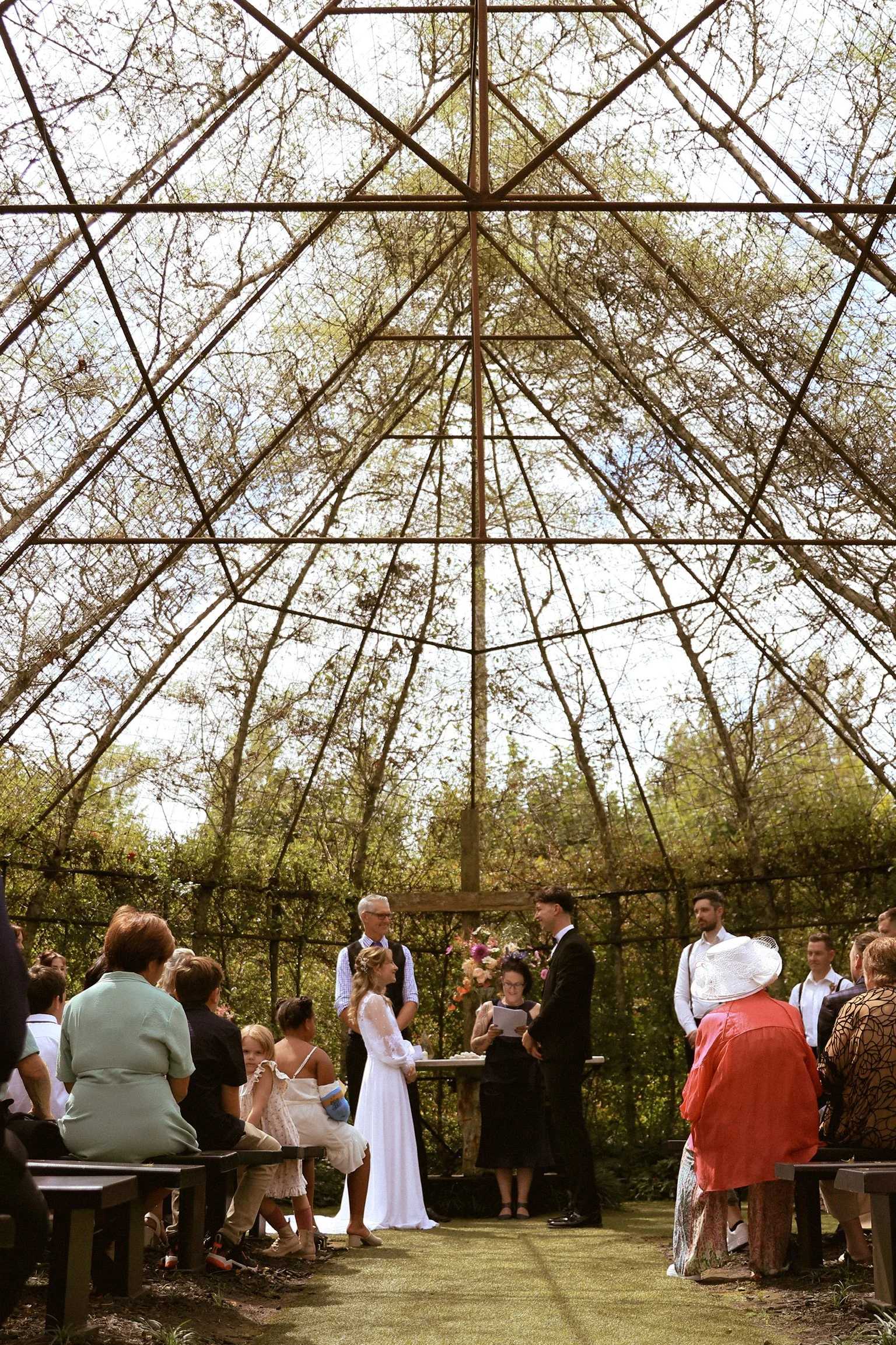 A wedding ceremony taking place outdoors inside a glass greenhouse, with a bride and groom standing before an officiant, surrounded by seated guests and a rustic, natural setting with green trees. Wedding Photography Waikato New Zealand. 