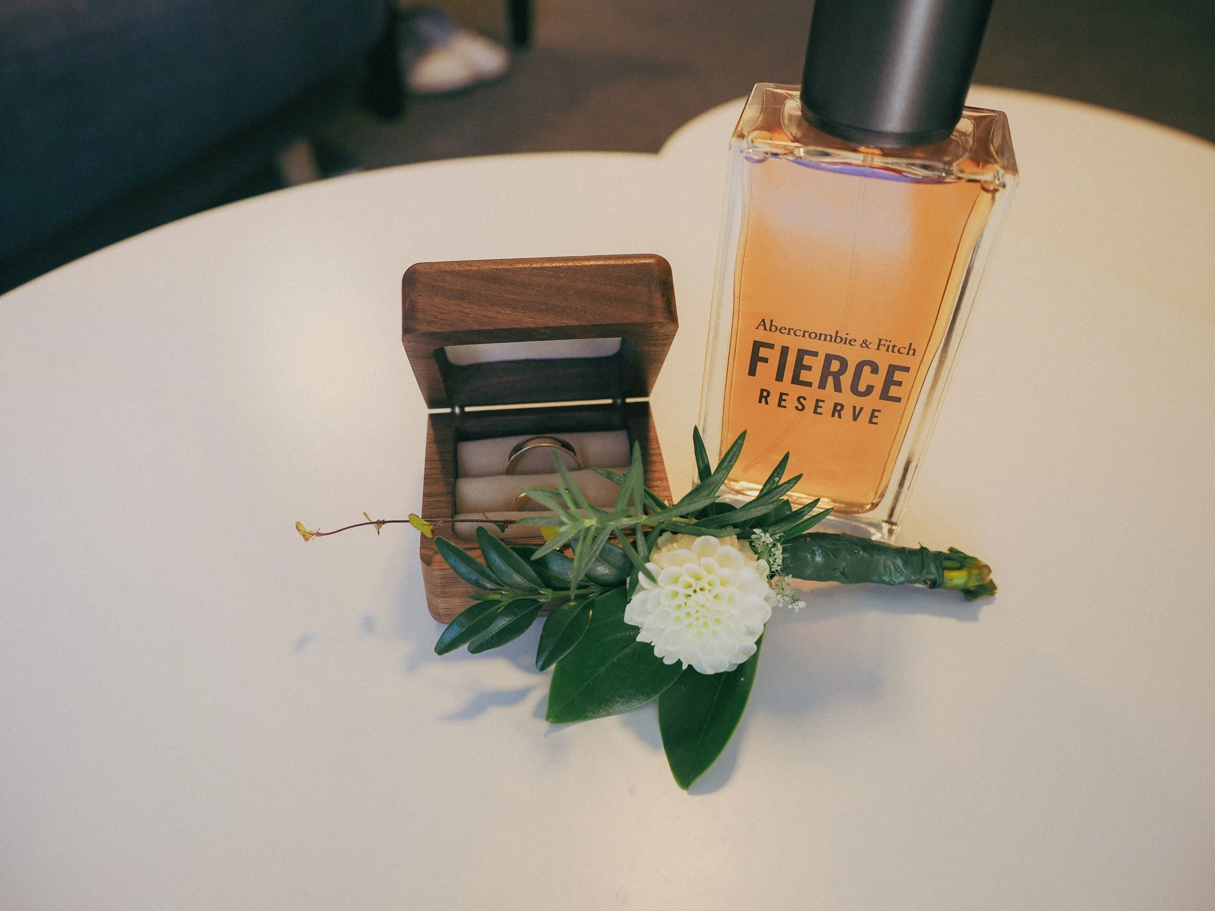 A bottle of Abercrombie & Fitch Fierce Reserve perfume placed on a white surface alongside a small wooden box with a ring inside and decorated with green leaves and white flowers.
