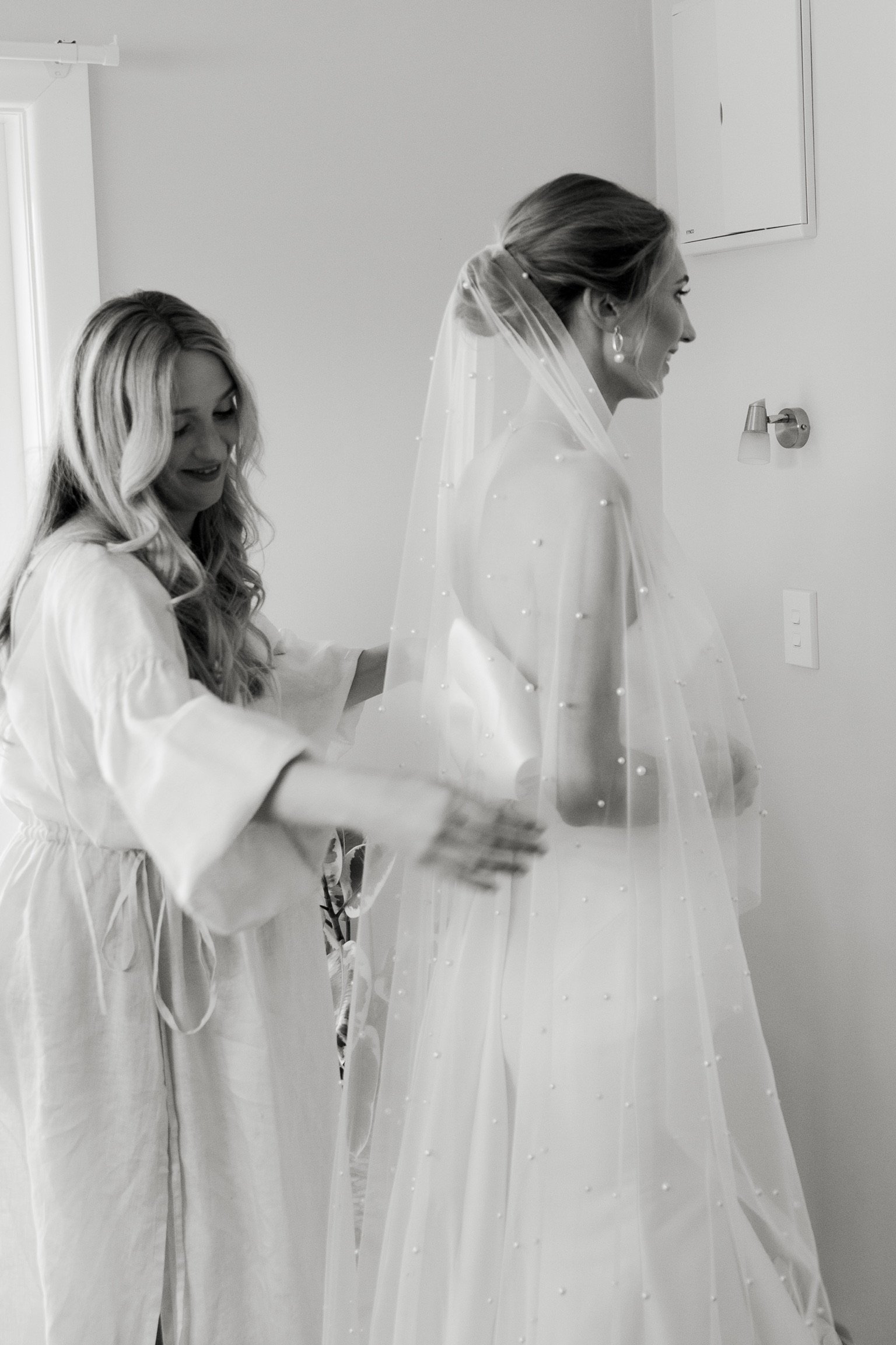 A bride in a wedding dress and veil smiling as a woman helps her get ready, possibly on her wedding day.