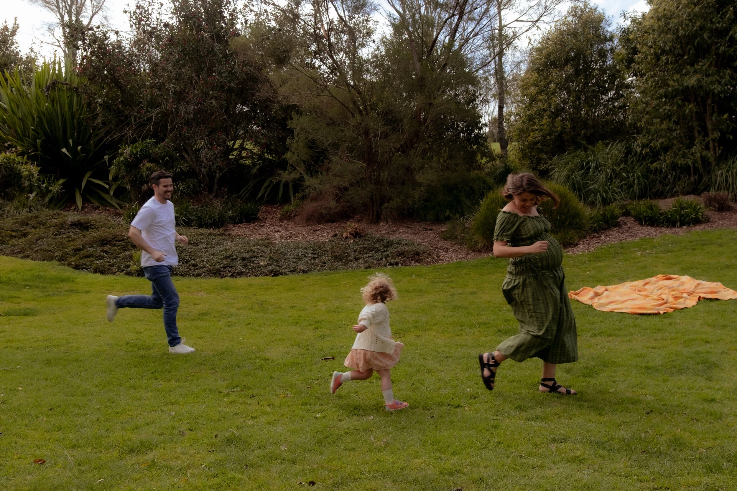 Family of three: a man, a woman, and a young girl, running on a grassy field outdoors. The woman is pregnant, and all are smiling and enjoying a playful moment. There is a rolled-up blanket on the ground in the background, with trees and bushes behin