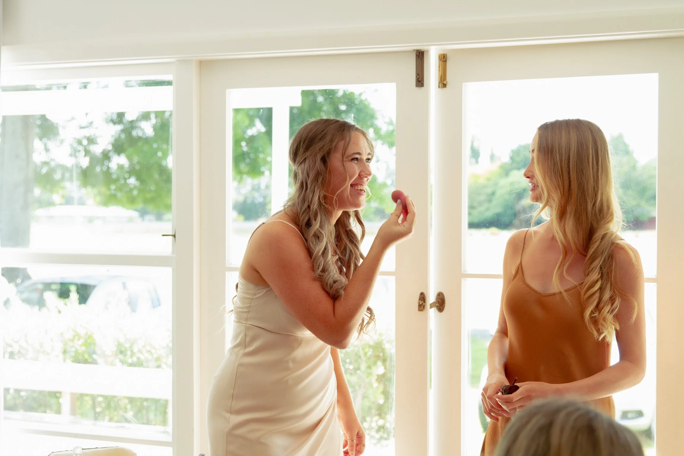 Two women are standing inside near large windows, smiling and engaging in conversation, one appears to be showing a small object in her hand.  Wedding Photography Waikato New Zealand. Wedding Photography Hamilton New Zealand. Northland Wedding Photo