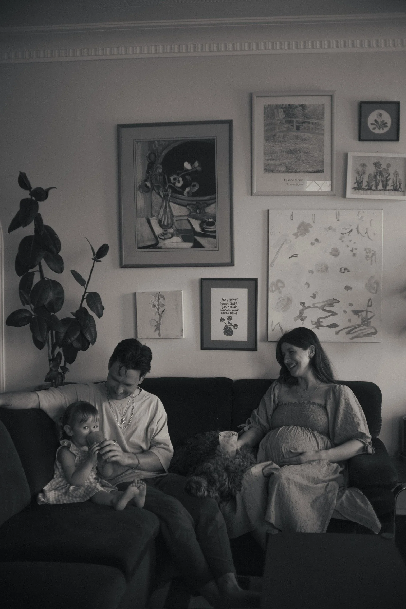 A black and white photo of three people and a dog sitting on a couch in a living room. The girl on the left is drinking, the man in the middle is smiling at the girl, and the woman on the right, pregnant, is smiling. Family Photographer. Hamilton