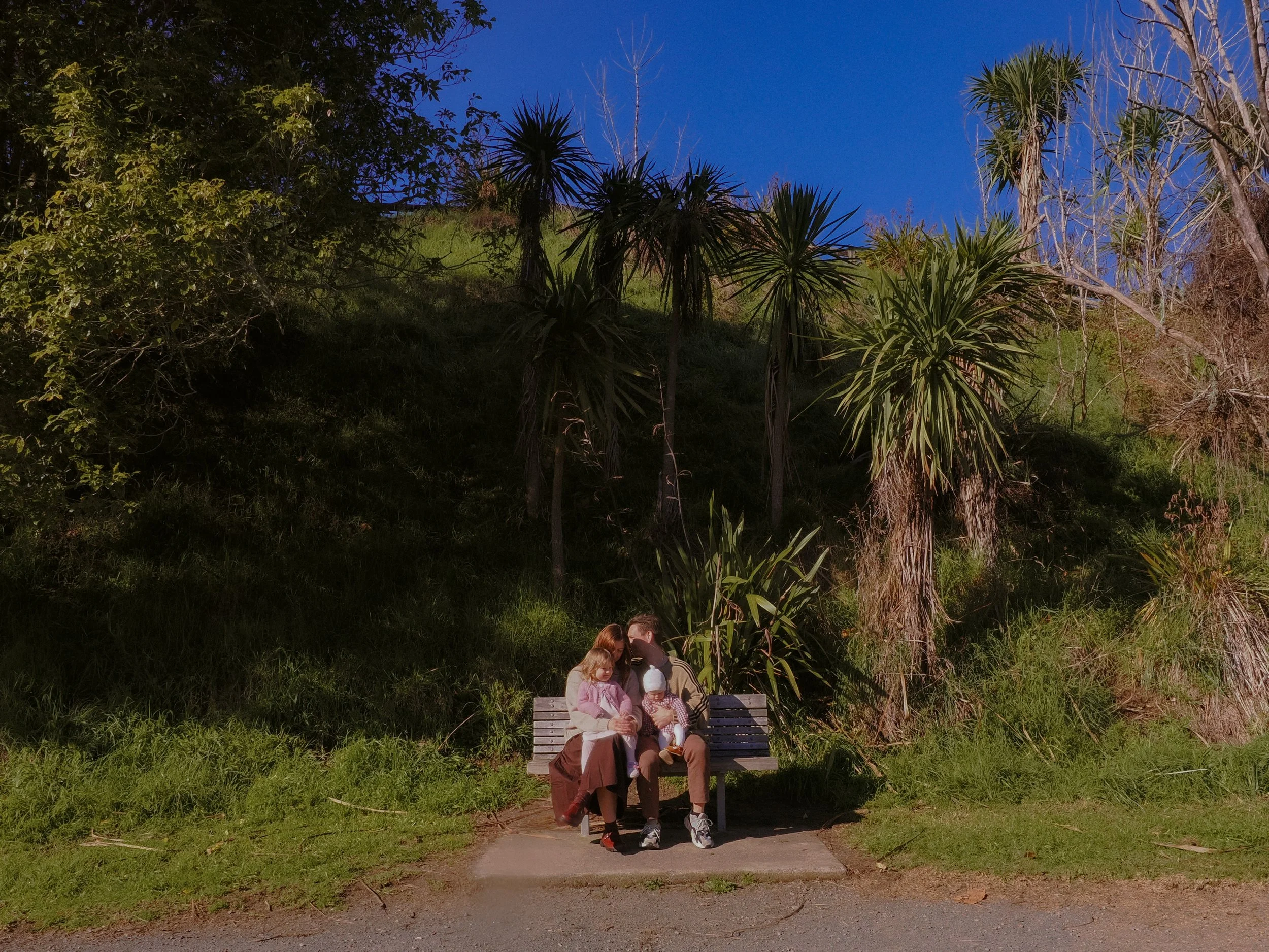 A family of three sitting on a bench in a park, with tall trees and green grass surrounding them, under a clear blue sky. Family Photography Waikato New Zealand. Family Photography Hamilton New Zealand. Northland Wedding Photography