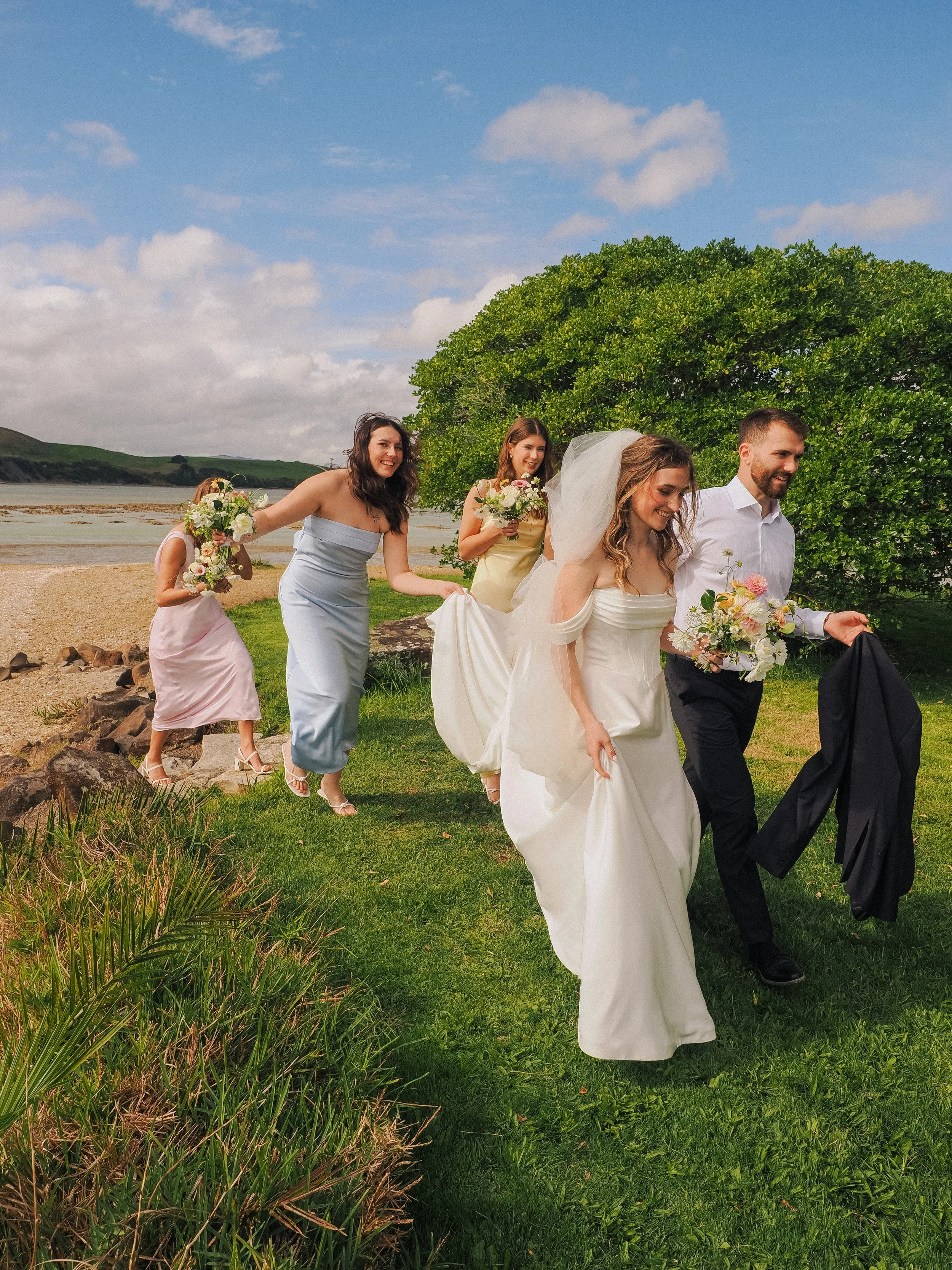 A wedding party walking outdoors on grass near a body of water, with a large green tree in the background, under a partly cloudy sky.  Wedding Photography Waikato New Zealand. Wedding Photography Hamilton New Zealand. Northland Wedding Photography