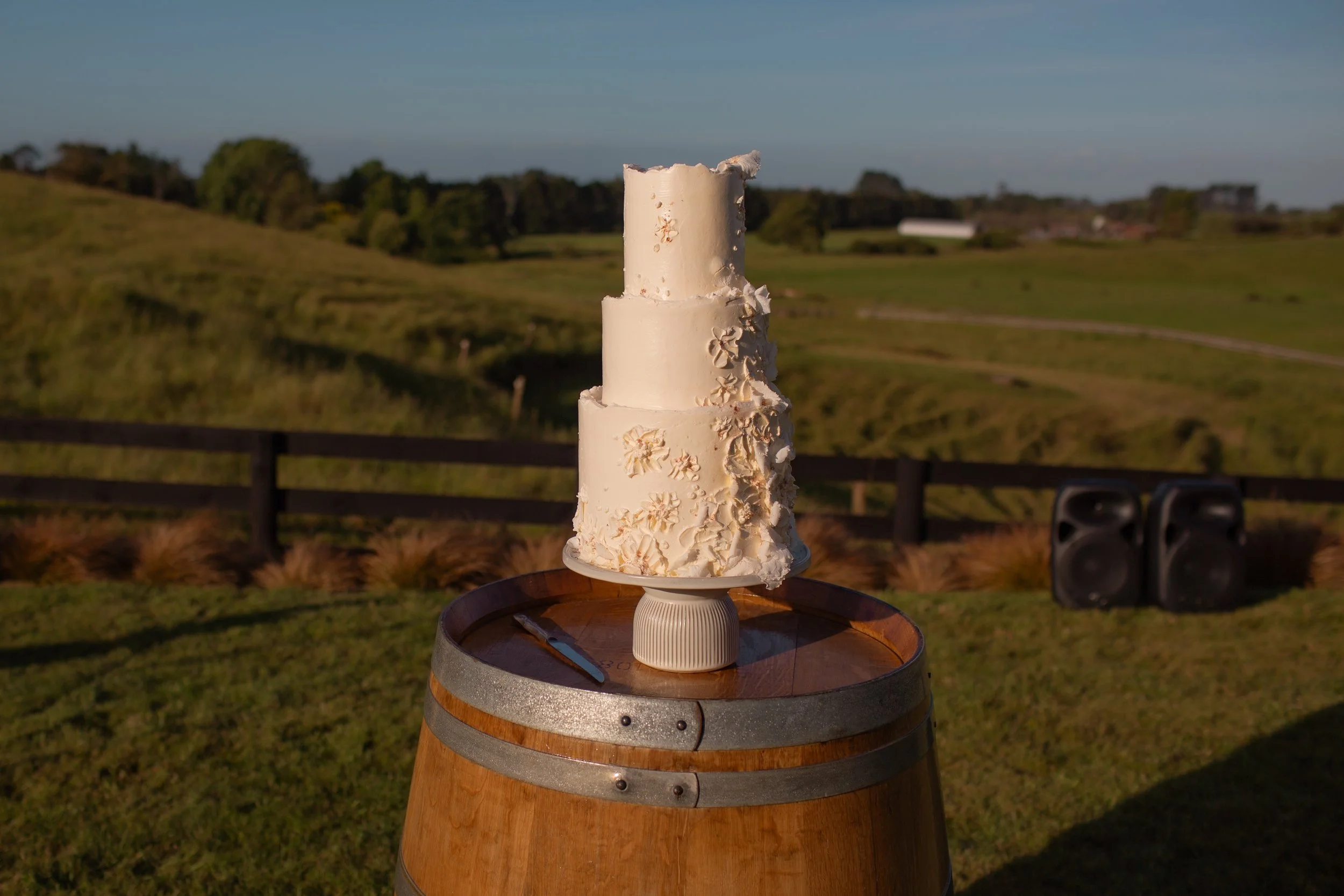 A multi-tiered white wedding cake with floral decorations sitting on top of a wooden barrel outdoors at sunset, with a hill, fence, and trees in the background.  Wedding Photography Waikato New Zealand. Wedding Photography Hamilton New Zealand. 