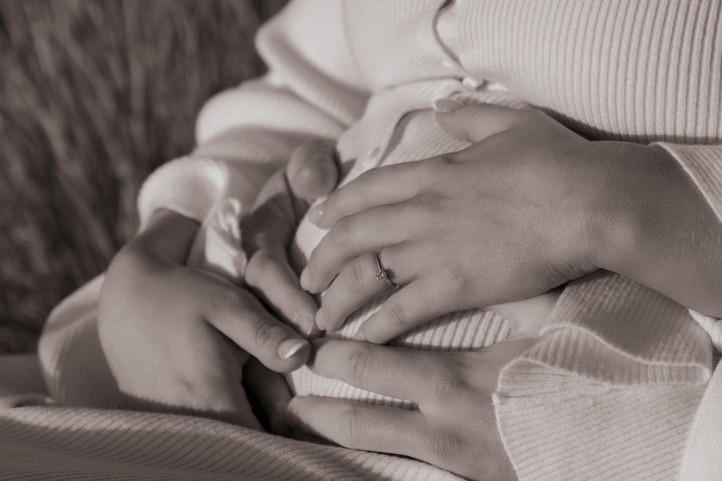 Two hands, one with an engagement ring, are gently holding each other on a person's lap, dressed in a long sleeve shirt. Maternity Shoot. Raglan New Zealand