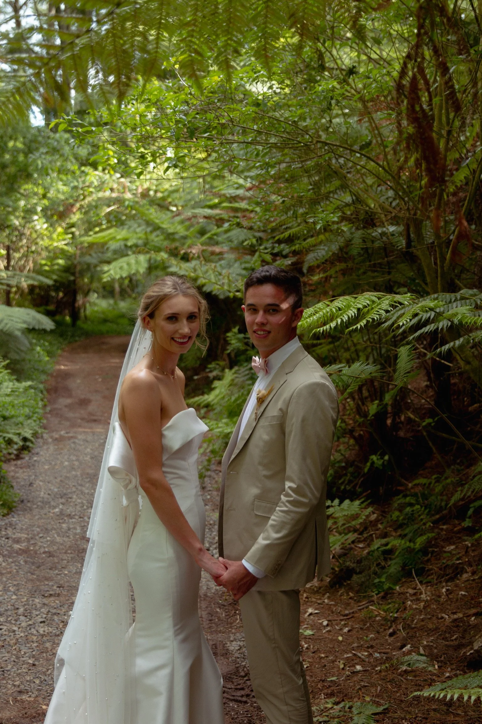 A newlywed couple holding hands in a lush forest, smiling at the camera. The bride is in a white strapless wedding gown with a veil, and the groom is in a light-colored suit with a bow tie.  Wedding Photography Waikato New Zealand. Wedding Photograph