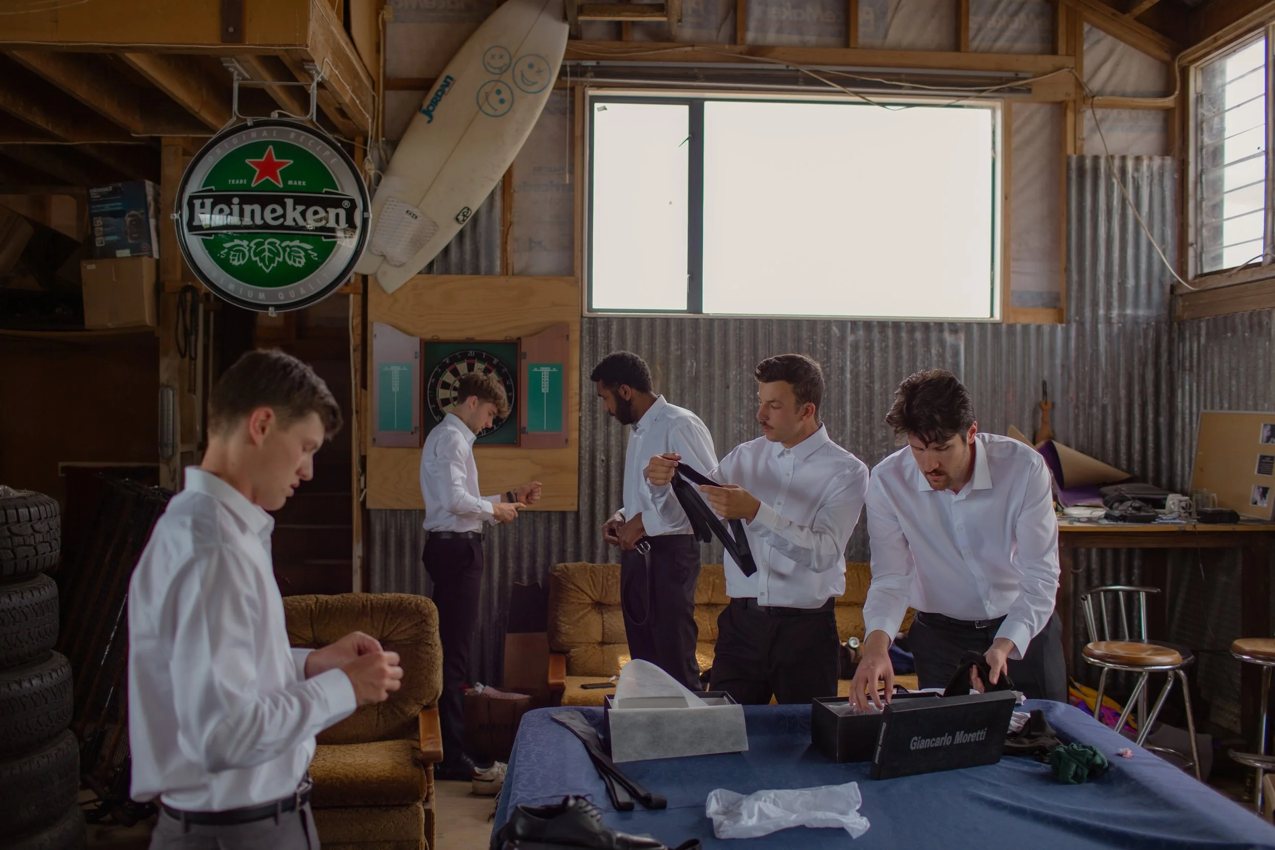 Five men in white shirts in a garage or workshop, preparing or packing black ties. The space has stacked tires, a Heineken sign, and various tools and items, with a large window letting in natural light.
