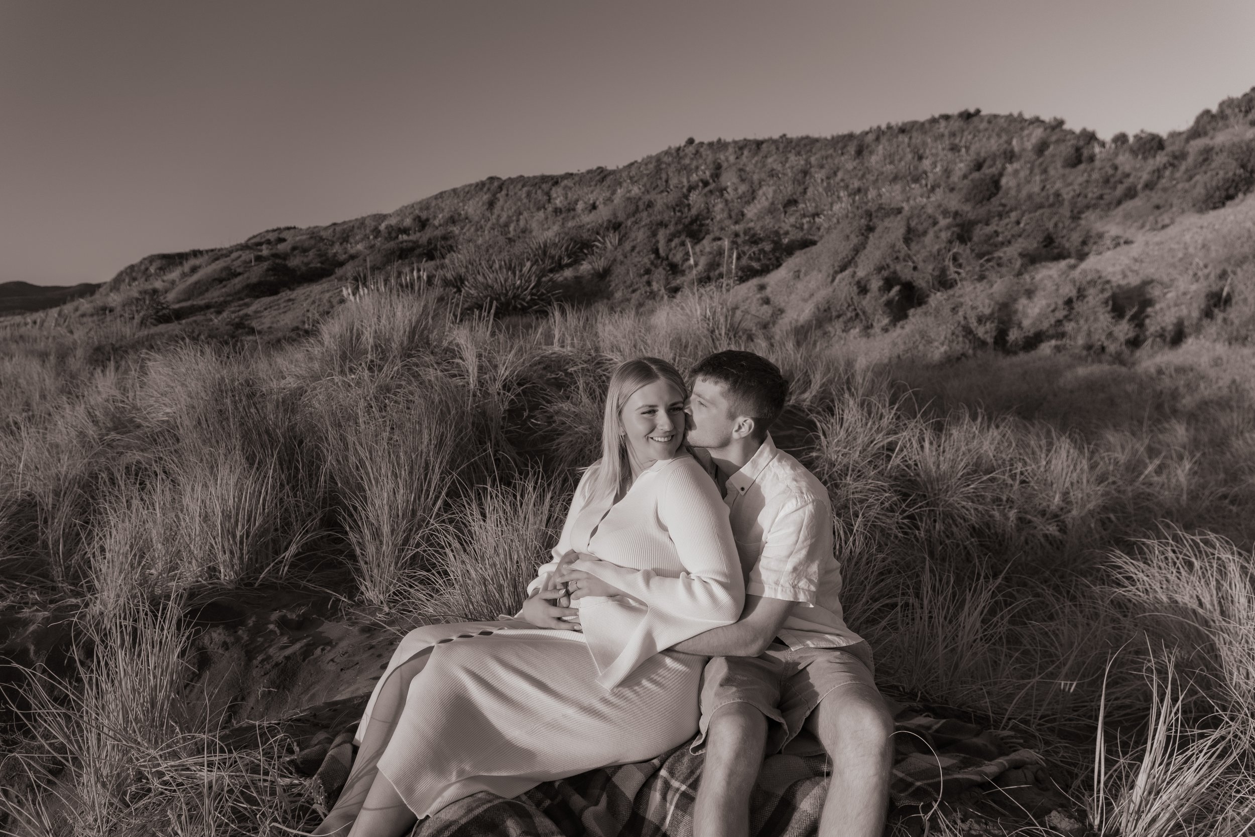 A black and white photo of a couple sitting on a blanket in a grassy outdoor area, with mountains in the background. The woman is smiling and facing the camera while the man kisses her cheek from the side. Maternity Shoot. Raglan New Zealand