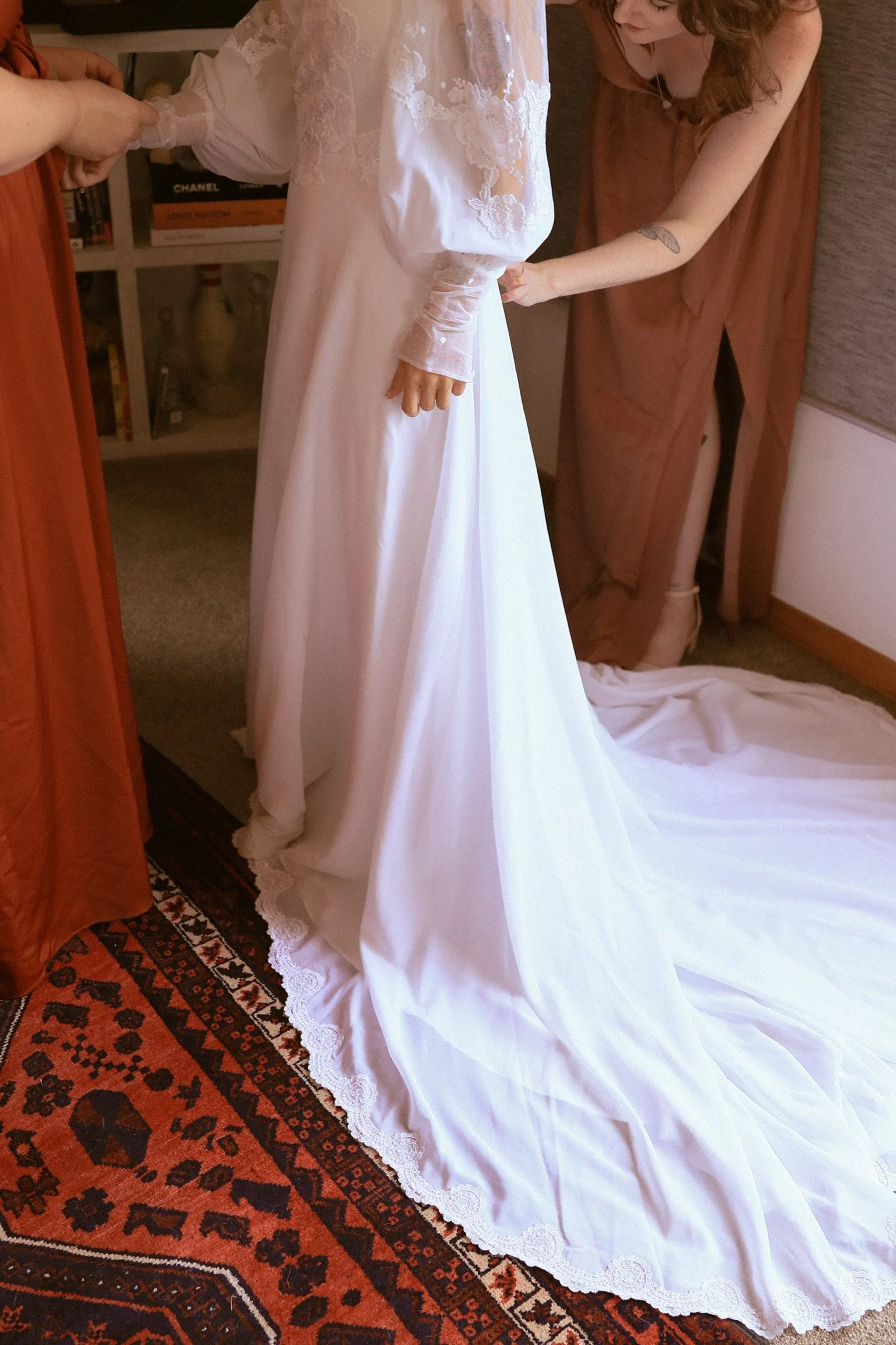 People adjusting a bride's white wedding gown with lace sleeves and a long train in a room with a patterned rug and a bookshelf. Wedding Photography Waikato New Zealand. Wedding Photography Hamilton New Zealand