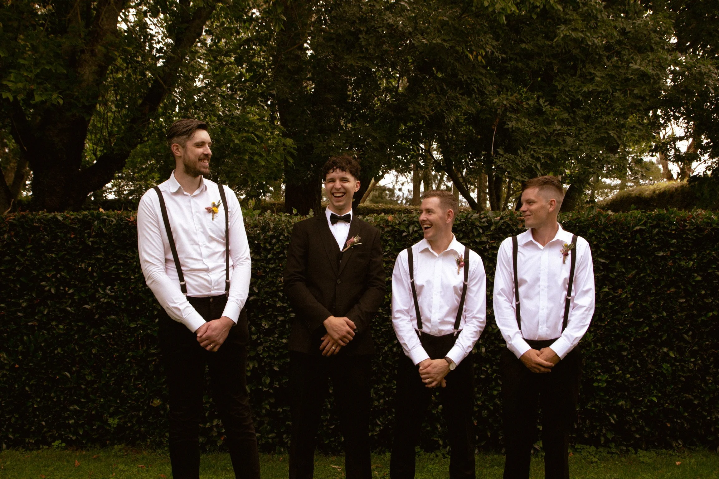 Four men in formal attire standing outdoors, smiling and laughing in front of a hedge with trees in the background. Wedding Photography Waikato New Zealand. Wedding Photography Hamilton New Zealand