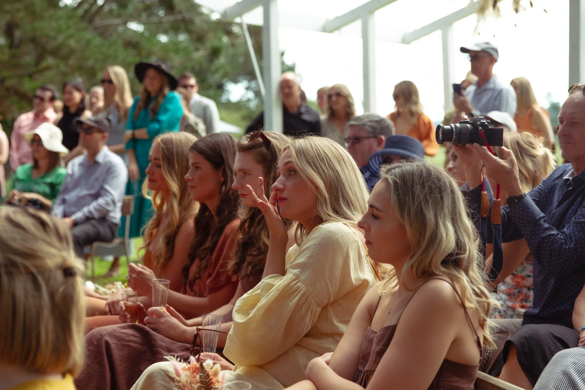 A group of people seated and standing outdoors under a canopy, watching an event. Some are taking photos, and others are observing attentively.  Wedding Photography Waikato New Zealand. Wedding Photography Hamilton New Zealand. Northland Wedding 