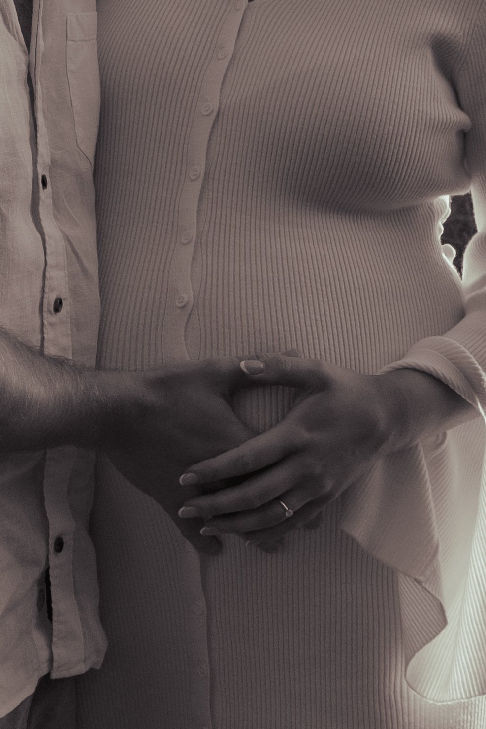Close-up of two people’s hands clasped together, one wearing a wedding ring, with one person in a buttoned shirt and the other in a ribbed long-sleeve top. Maternity Shoot. Raglan New Zealand
