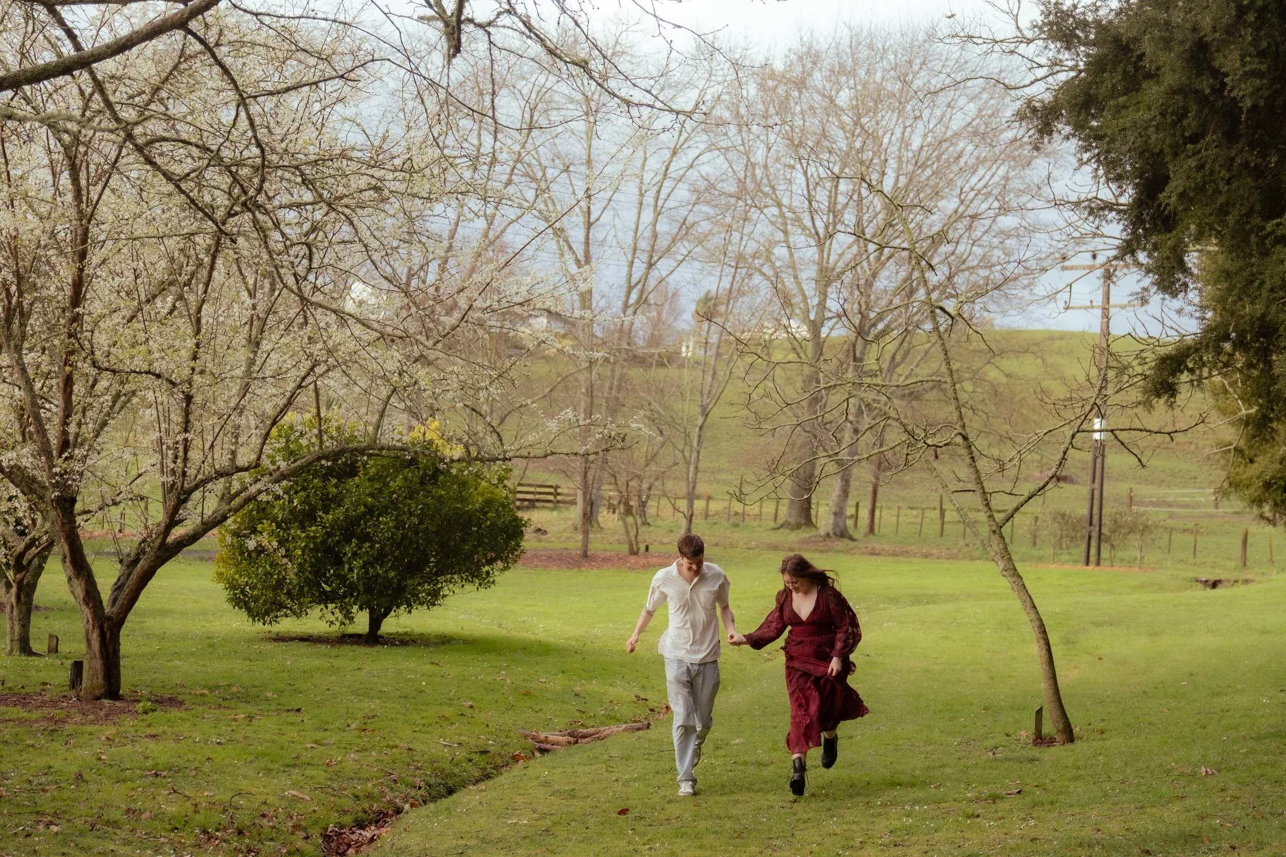 A young couple holding hands and running together on a grassy path in a park-like area with trees and a hill in the background. Couples photography Raglan New Zealand. Engagement photography Hamilton New Zealand. Whangarei New Zealand Photographer