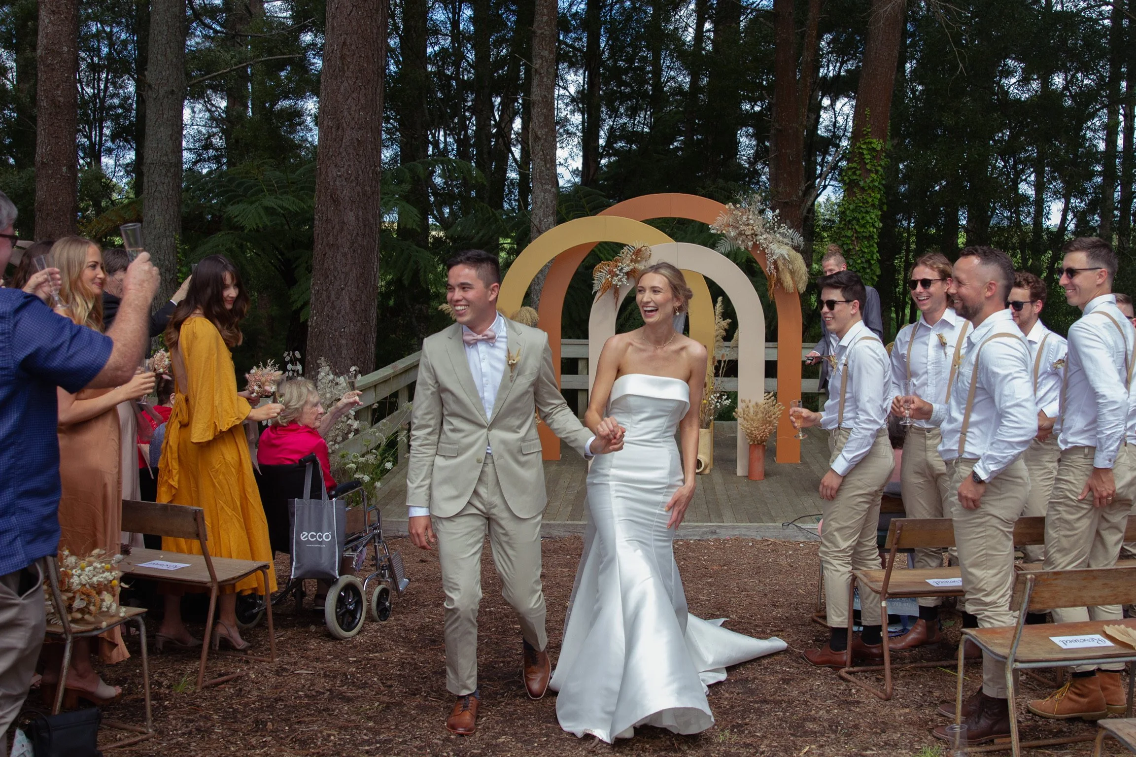 Bride and groom holding hands and smiling during outdoor wedding ceremony in a forest, with guests and decorative arch behind them.  Wedding Photography Waikato New Zealand. Wedding Photography Hamilton New Zealand. Northland Wedding Photography