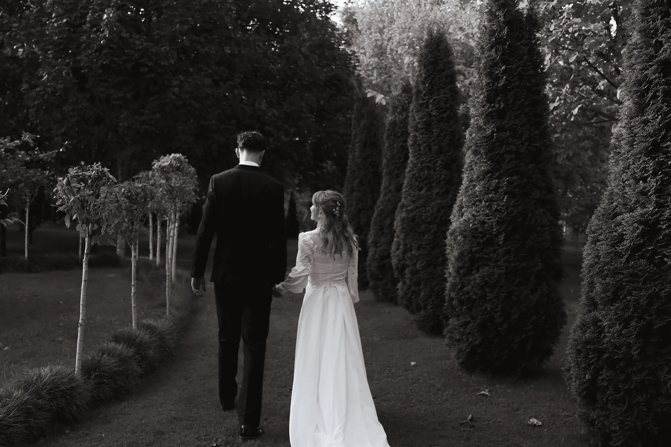 Black and white photo of a bride and groom walking hand in hand on a garden path with tall trees on each side. Wedding Photography Waikato New Zealand. Wedding Photography Hamilton New Zealand