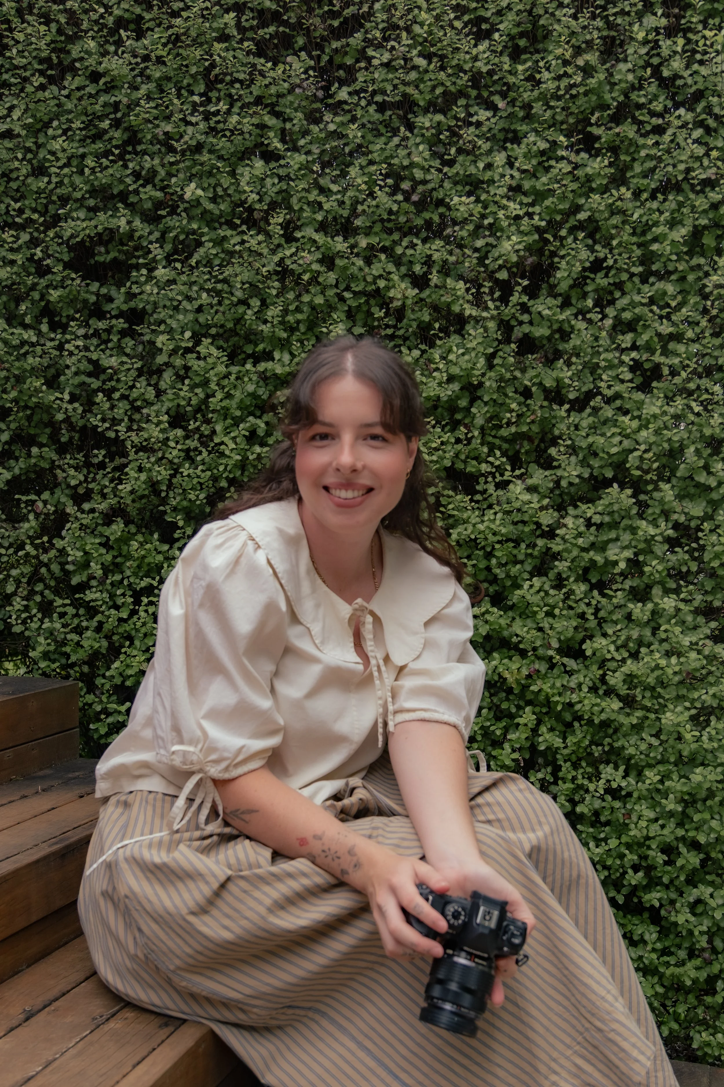 A woman with brown hair, wearing a cream-colored blouse and striped pants, sitting on a wooden bench in front of a green leafy wall, holding a camera in her hands and smiling.