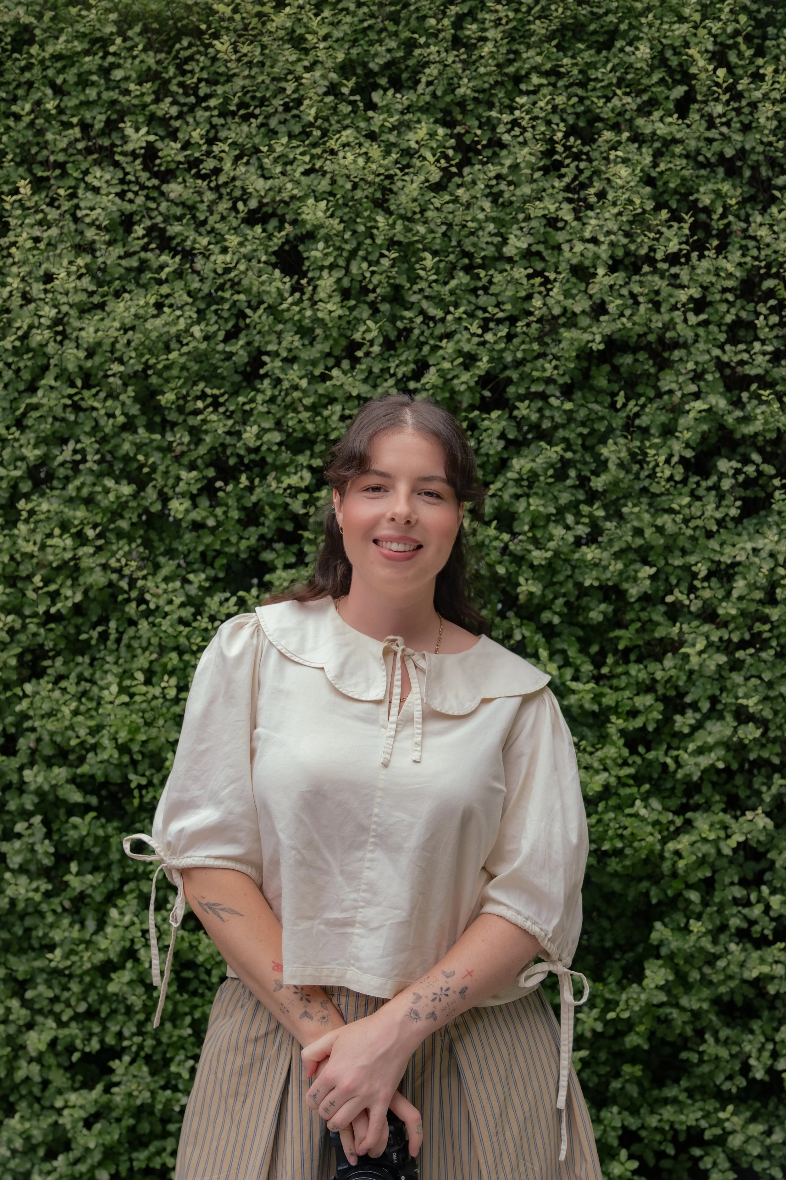 A woman with brown hair and tattoos on her forearms standing in front of a green leafy hedge, smiling at the camera. She is wearing a light-colored blouse with a ruffle collar and striped pants, holding a camera in her hands.