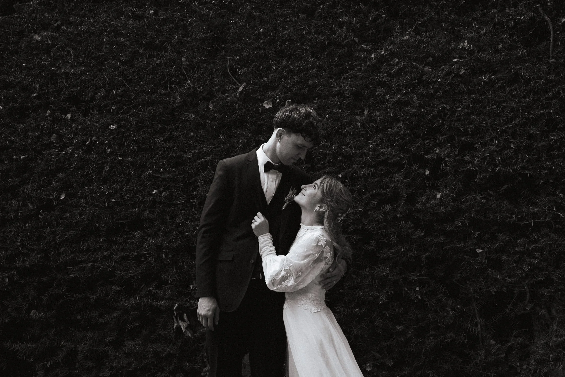 A black and white photo of a bride and groom standing close, looking into each other's eyes, with a dark leafy background. The groom wears a tuxedo with a bow tie, and the bride wears a long lace wedding dress, holding a small bouquet.