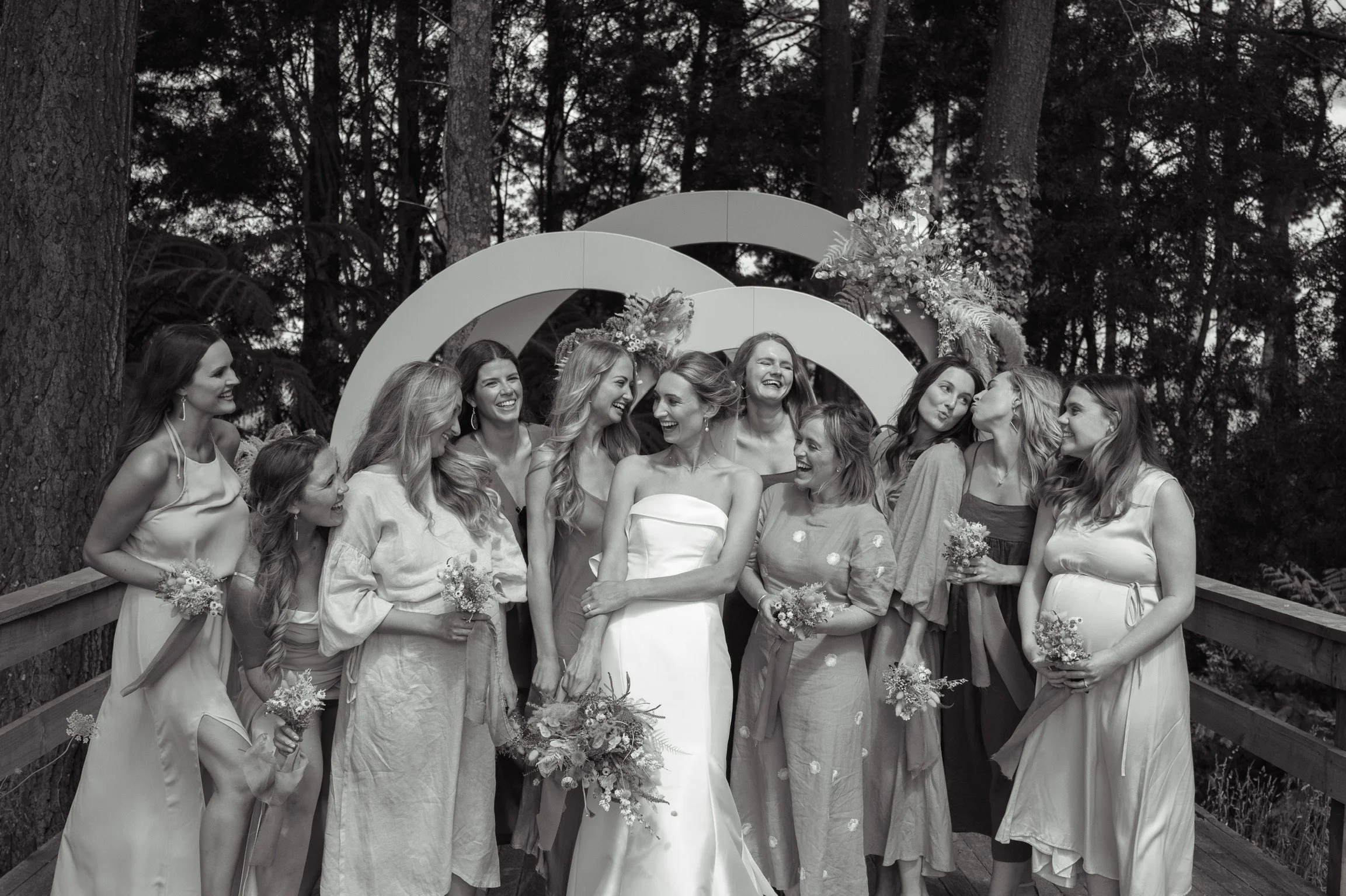 Black and white photo of a bride and twelve bridesmaids standing on a wooden bridge outdoors, with trees in the background. The bride is in the center holding a bouquet, and the bridesmaids are holding smaller bouquets, all smiling.