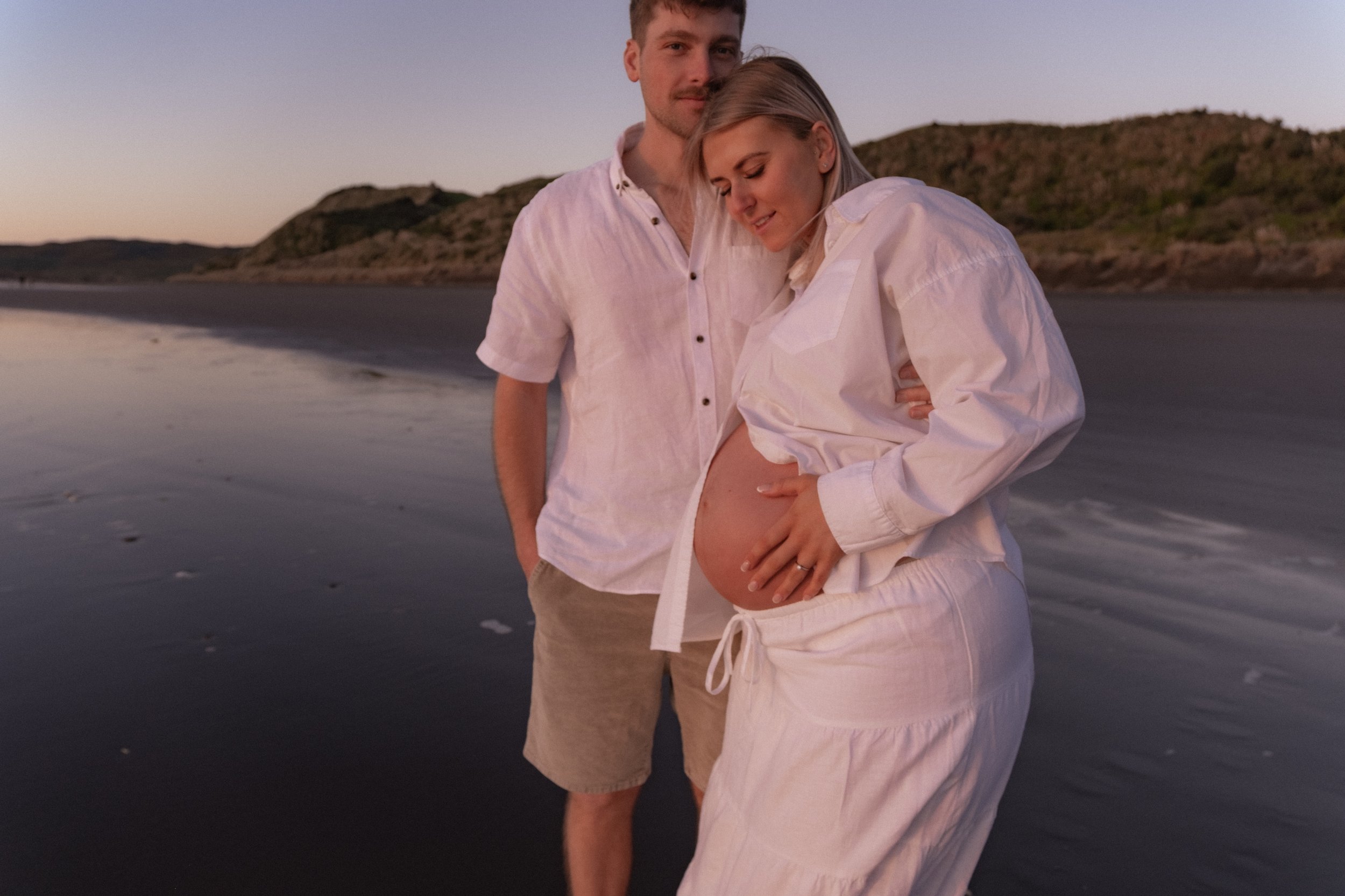 A pregnant woman and a man standing together on the beach at sunset, with the woman showing her belly and the man gently holding her from behind. Maternity photography. Raglan New Zealand Photographer