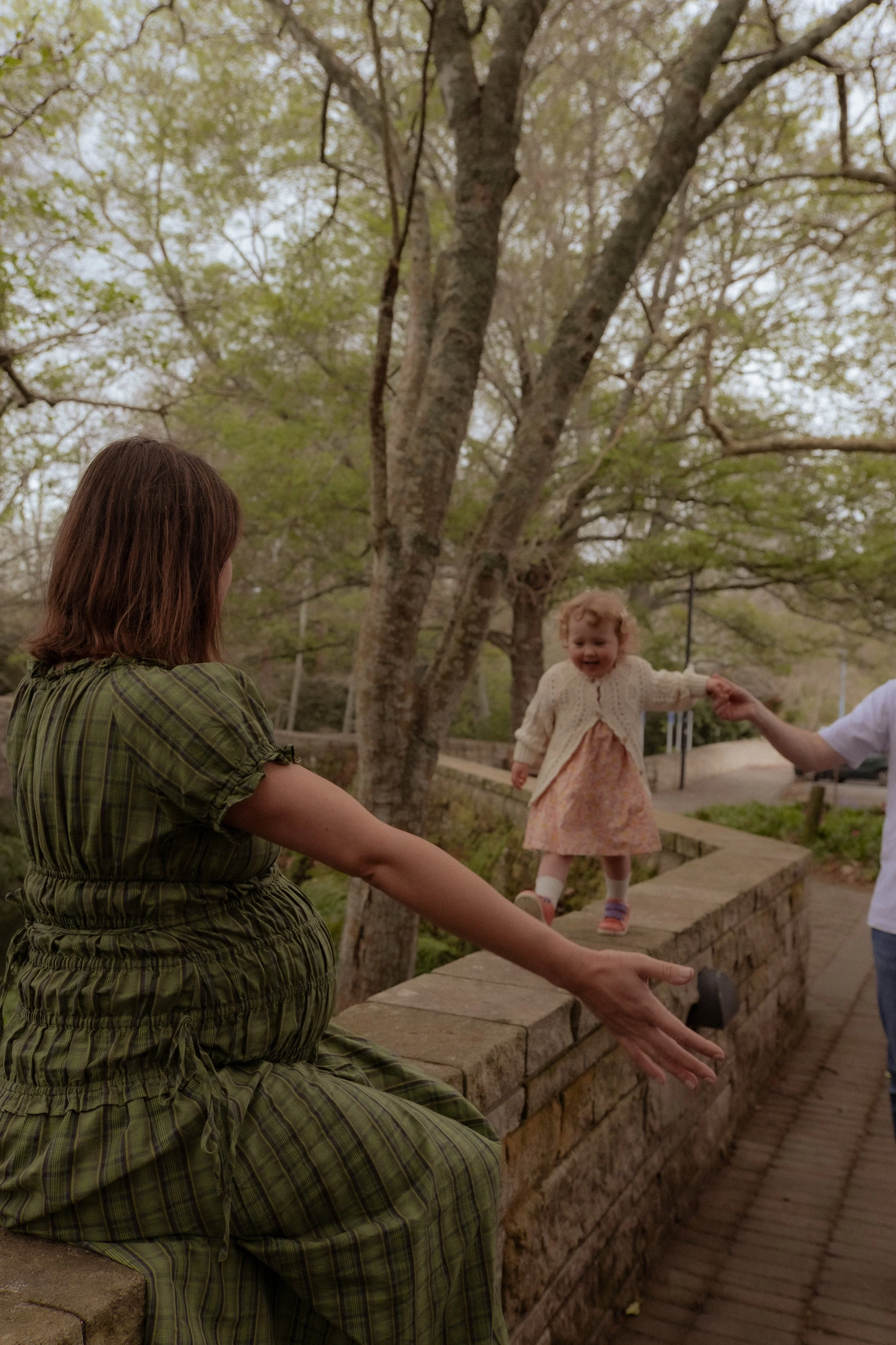 A woman in a green plaid dress sitting on a brick wall, reaching out to a young girl in a pink dress and cream cardigan who is standing on the wall, holding hands with someone outside the frame amid trees with green leaves. Maternity photography