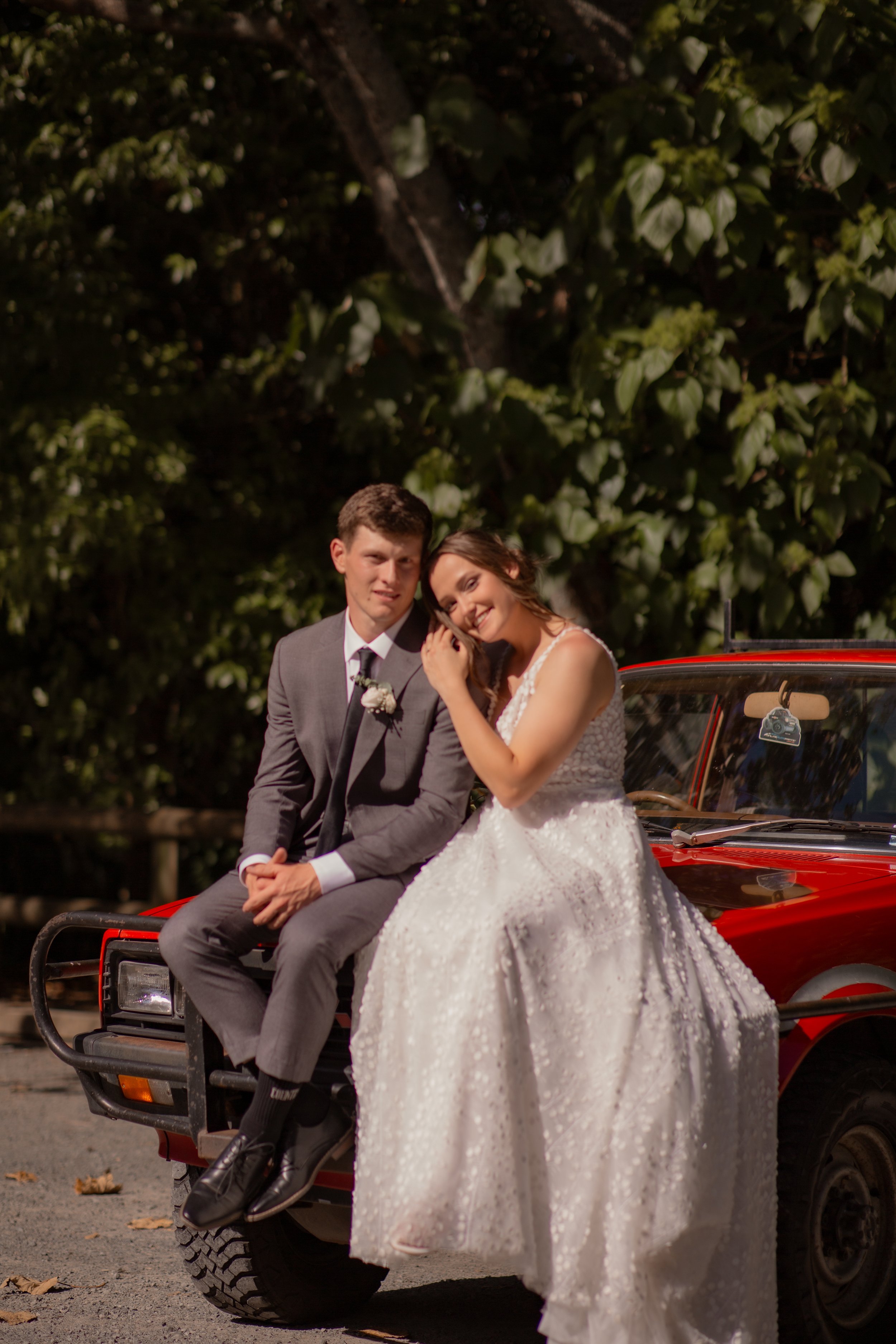 A bride and groom sitting on the hood of a red vintage car outdoors, with trees in the background. The bride is wearing a white wedding dress and smiling, while the groom is in a gray suit, white shirt, and black tie, looking at the camera.