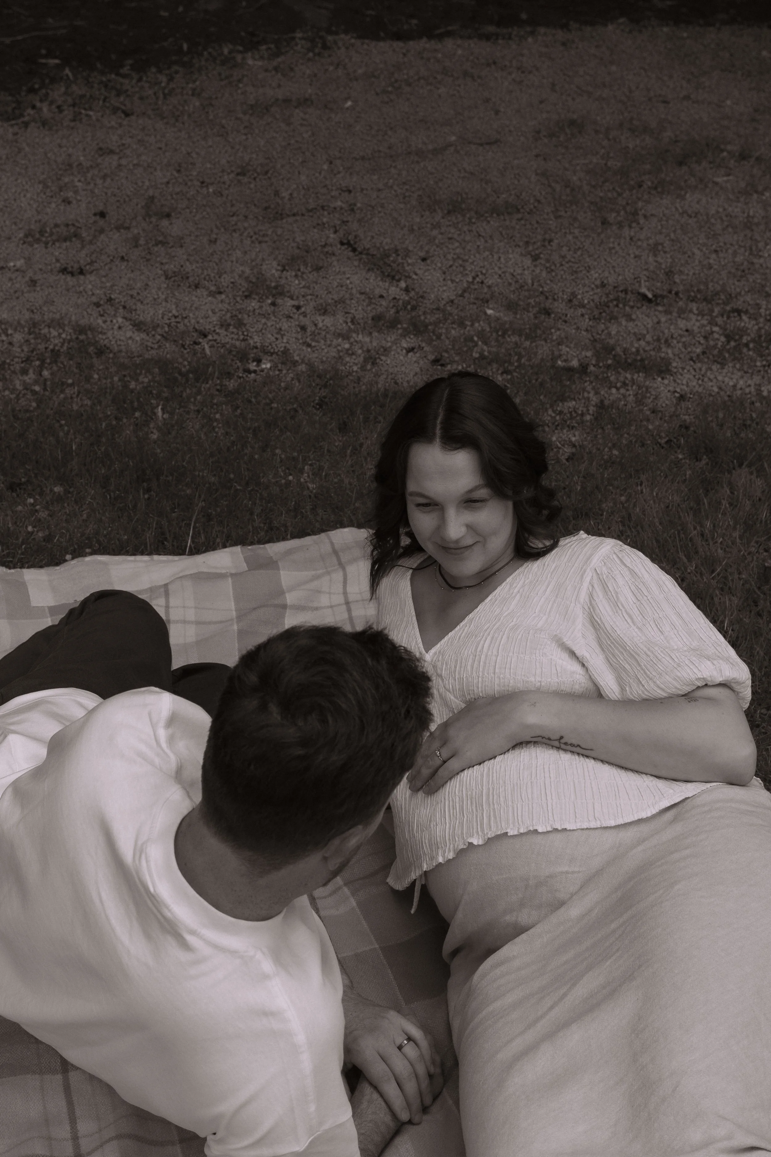 A woman lying on a blanket outside, looking at a man sitting beside her, with grass and dirt ground in the background. Family Photographer. Maternity Photography. Hamilton New Zealand Photographer