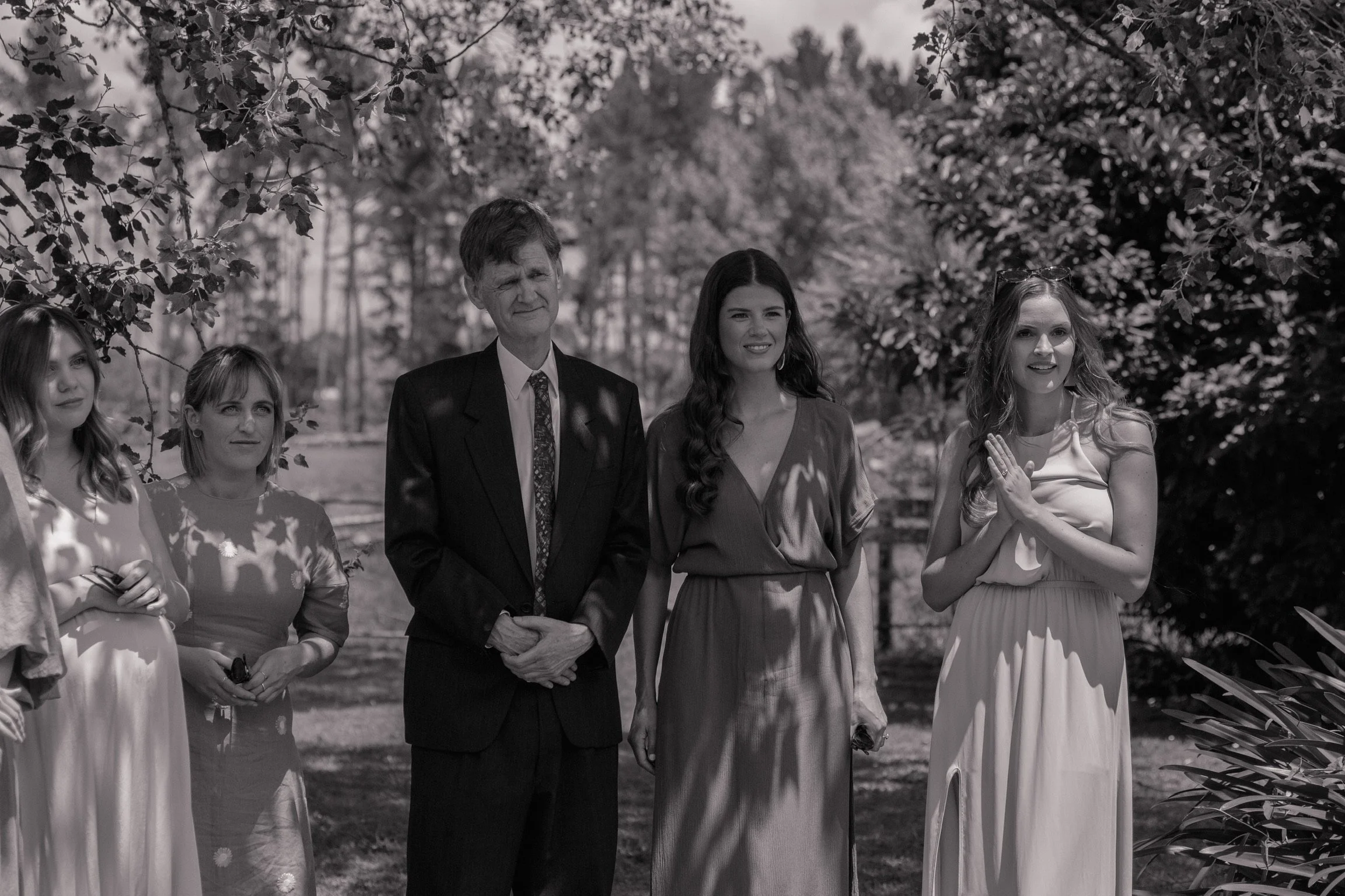 A black and white photo of five people standing in a garden surrounded by trees and plants, dressed in formal attire during daytime.  Wedding Photography Waikato New Zealand. Wedding Photography Hamilton New Zealand. Northland Wedding Photography