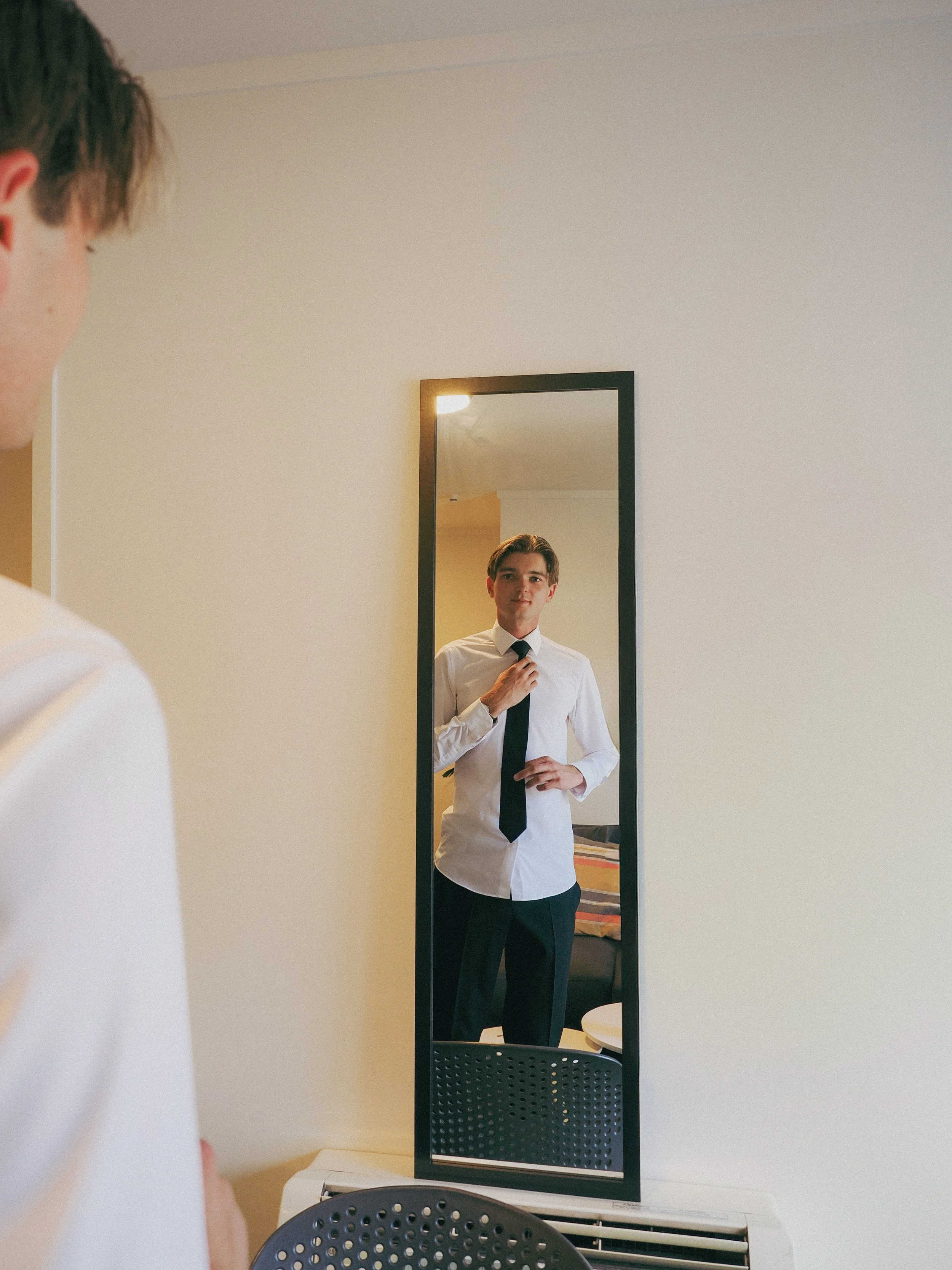 A man in a white shirt and black tie adjusting his tie while looking at himself in a full-length mirror in a room with a bed and a chair.