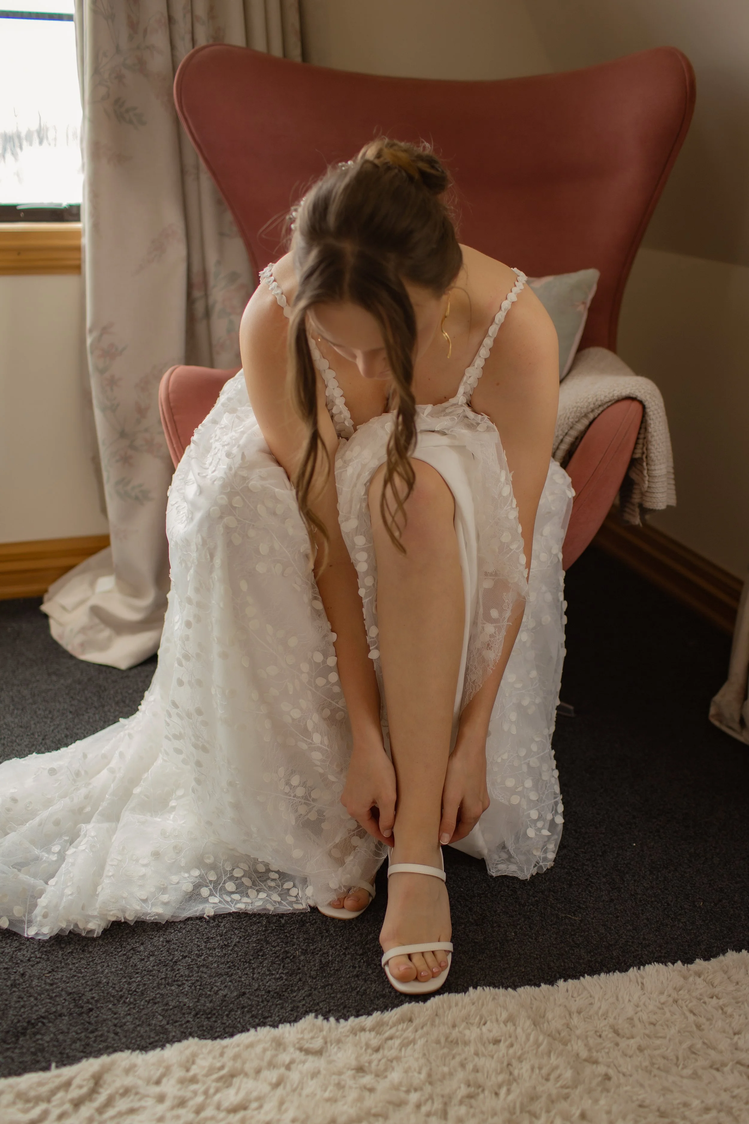 Bridal woman in a white dress adjusting her shoe while kneeling on a carpet in a cozy room with natural light.  Wedding Photography Waikato New Zealand. Wedding Photography Hamilton New Zealand. Northland Wedding Photography