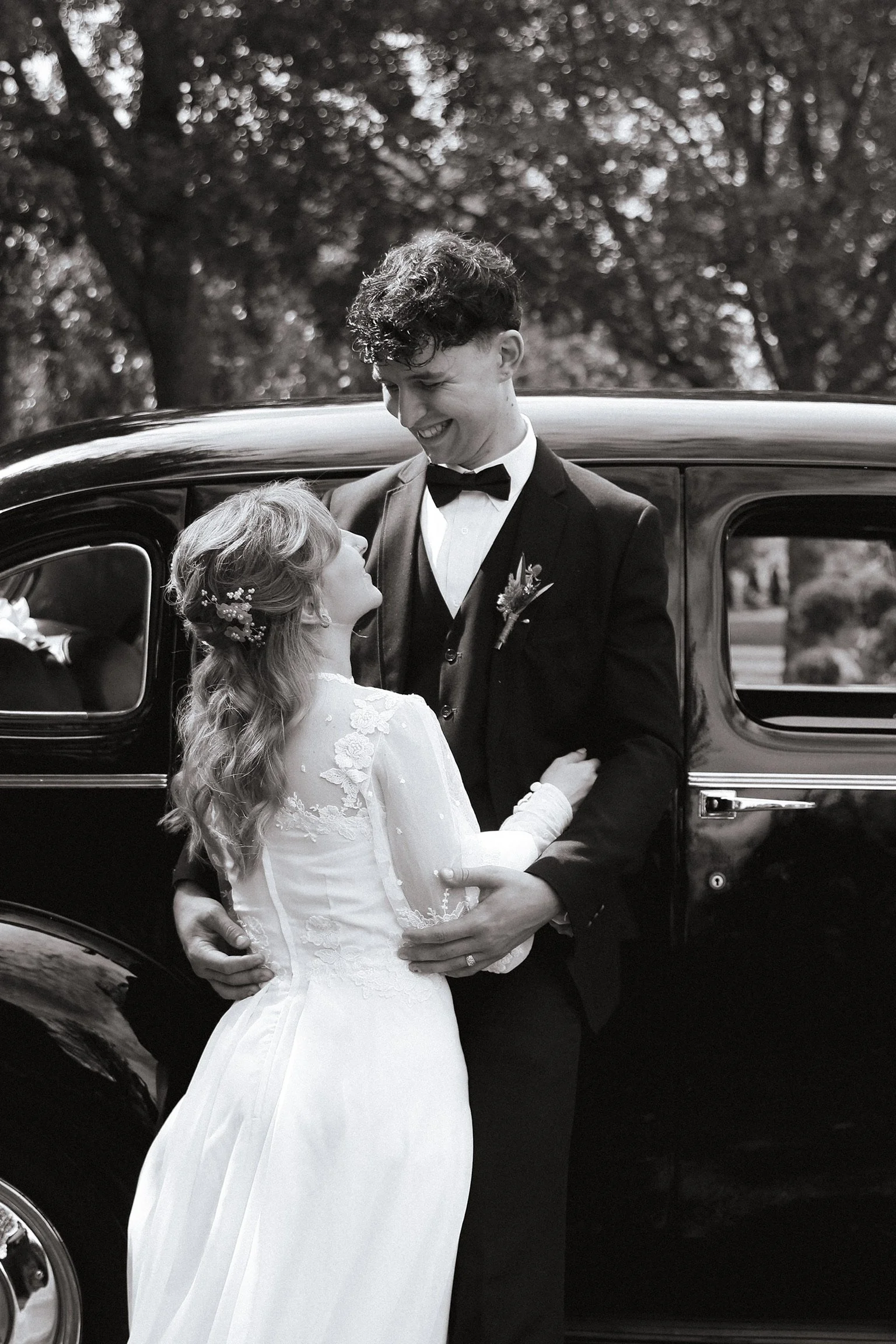 Black and white photo of a bride and groom standing in front of a vintage car, smiling and looking into each other's eyes. Wedding Photography Waikato New Zealand. Wedding Photography Hamilton New Zealand