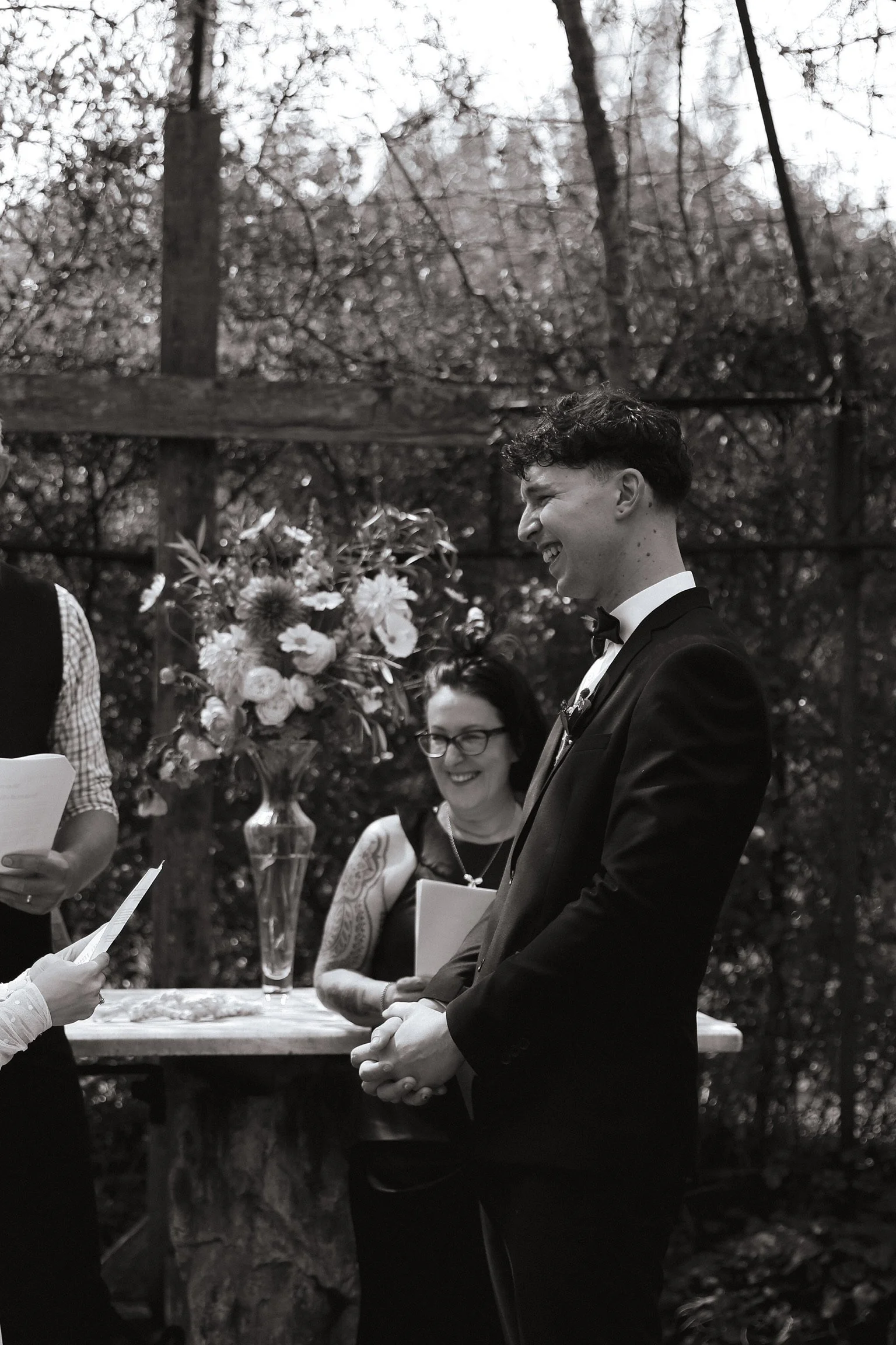 A black and white photo of a wedding ceremony outdoors with a smiling groom in a tuxedo and a woman with glasses and tattoos, with a large flower arrangement in a vase on a rustic table behind them. Wedding Photography Waikato New Zealand. 