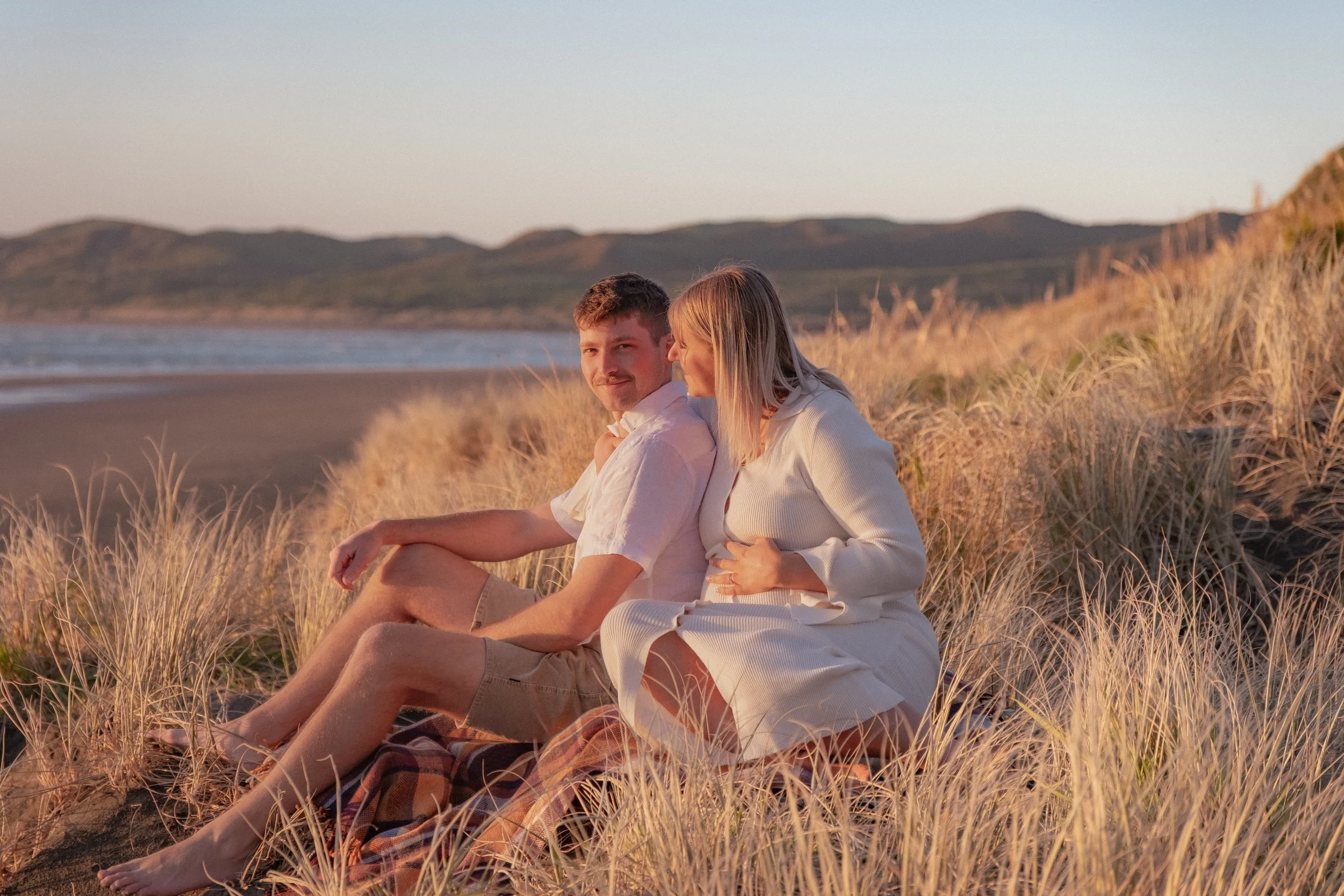 A couple sitting on a blanket on a sandy beach surrounded by tall grass, with the ocean and mountains in the background during sunset. Maternity Shoot. Raglan New Zealand