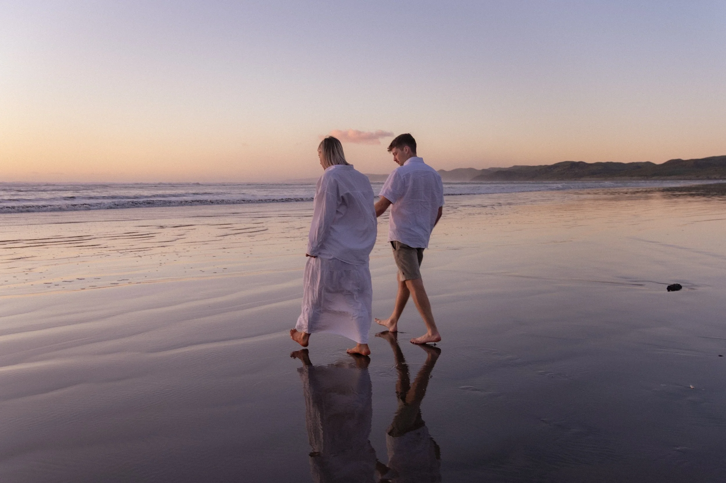 A couple walks along the beach at sunset, holding hands, with their reflections visible in the wet sand. Maternity Photography. Raglan New Zealand Photographer