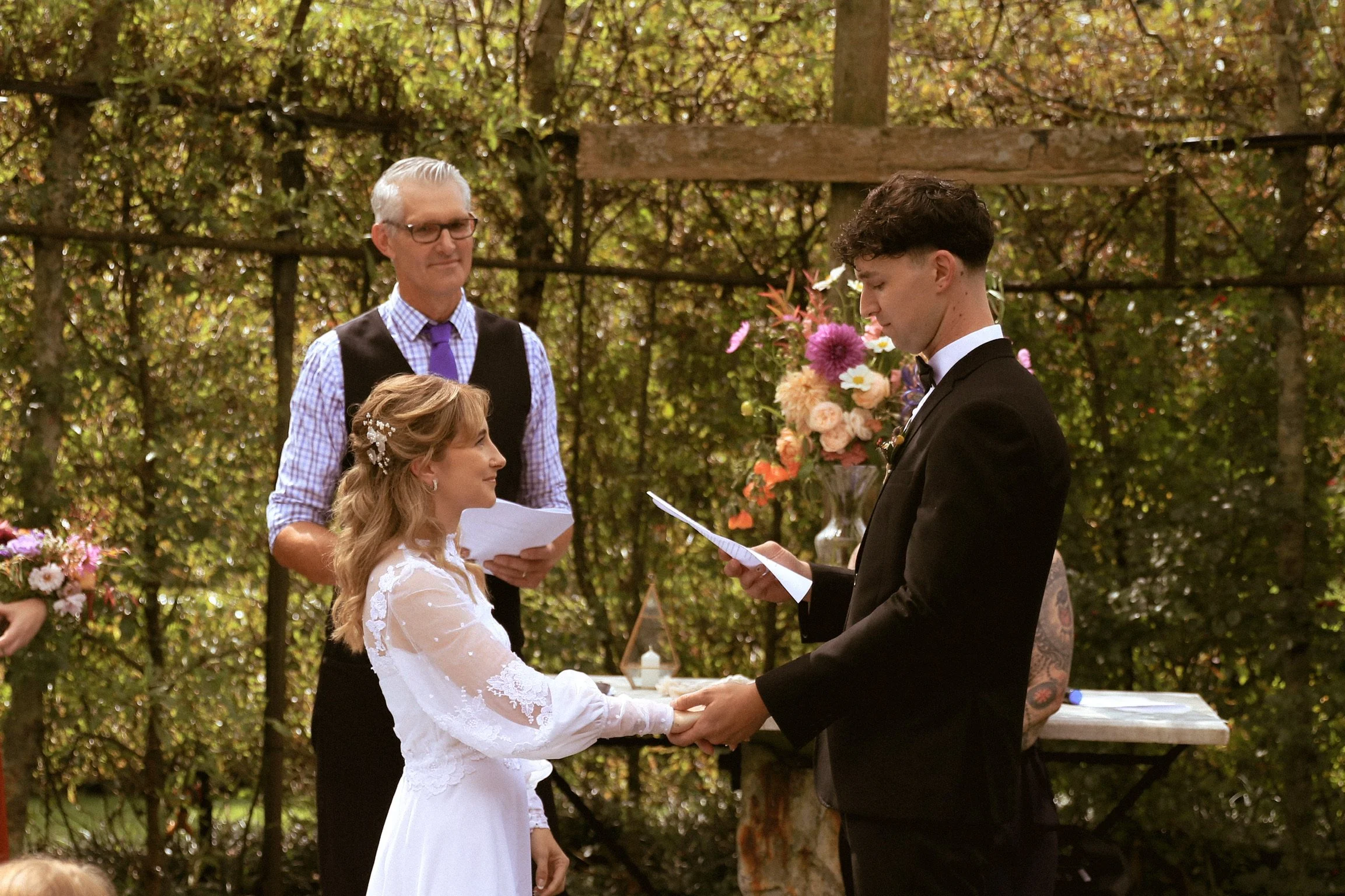 A couple getting married outdoors, holding hands and facing each other, with an officiant and a man reading vows in the background, surrounded by trees and flowers. Wedding Photography Waikato New Zealand. Wedding Photography Hamilton New Zealand