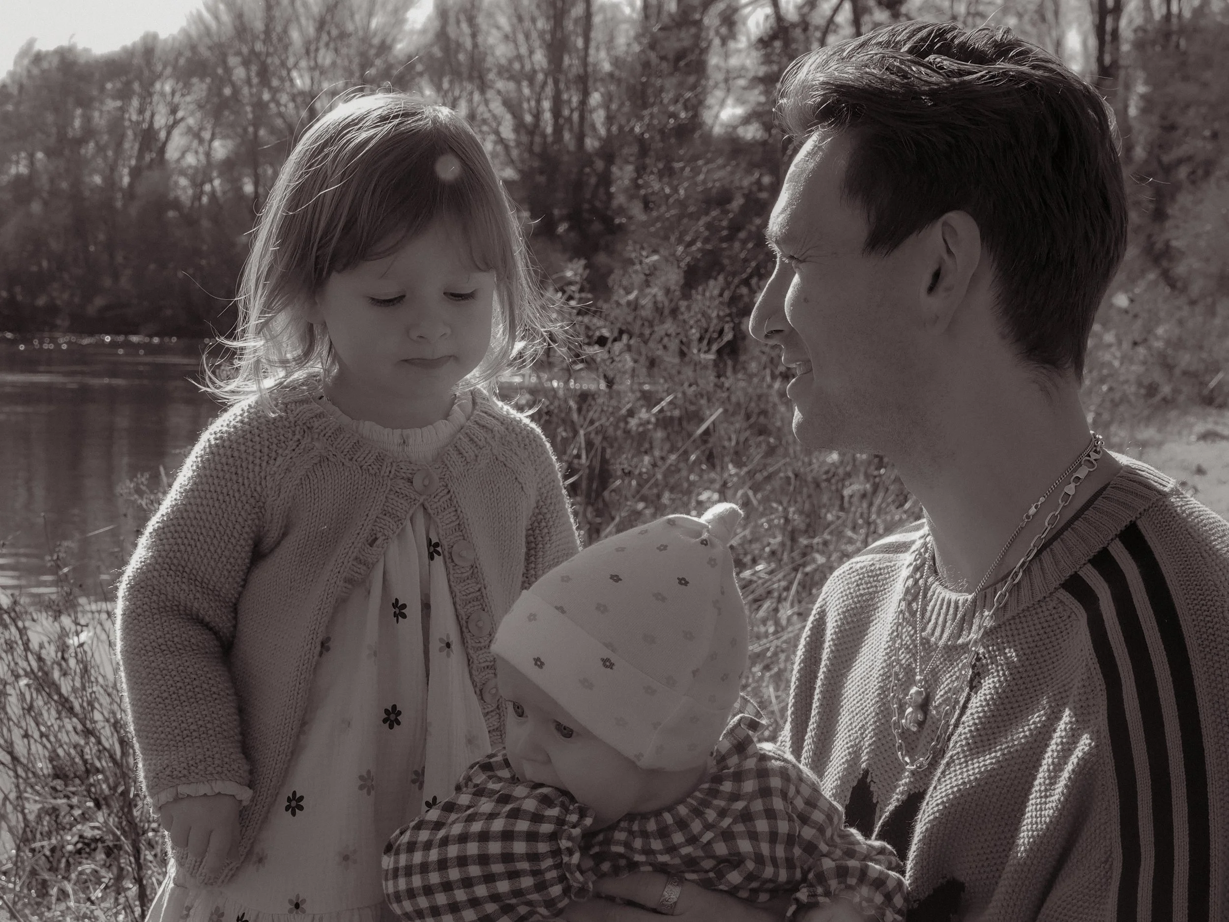 A man holding a baby girl wearing a checkered shirt and a hat, while a young girl in a dress and cardigan looks on beside them outdoors with a river and trees in the background. Family Photography Waikato New Zealand. Family Photography Hamilton 