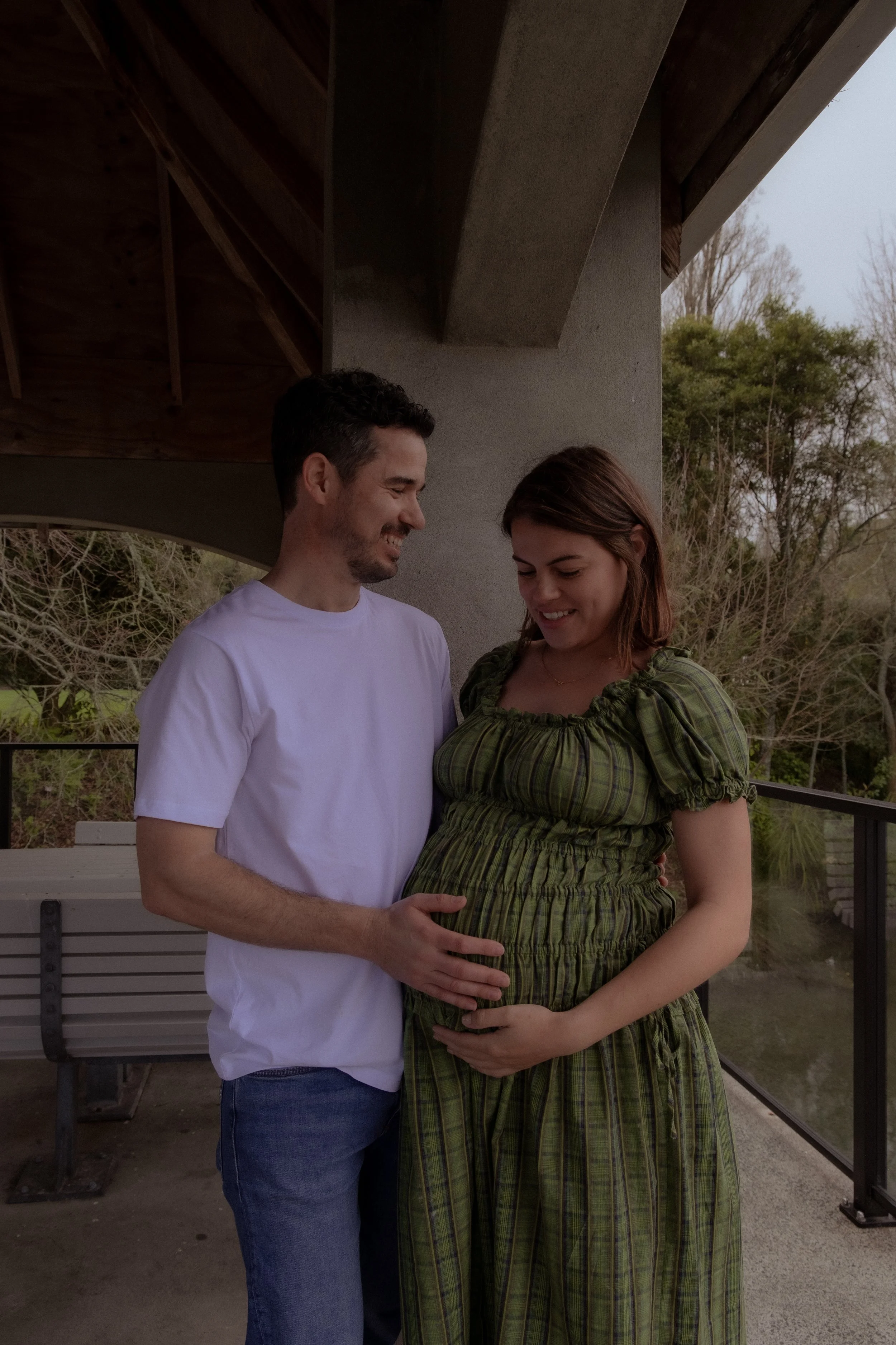 A happy couple pregnant woman and man standing outdoors on a balcony, the woman is touching her belly and both are smiling at each other. Maternity photography Hamilton New Zealand. Whangarei New Zealand Photographer