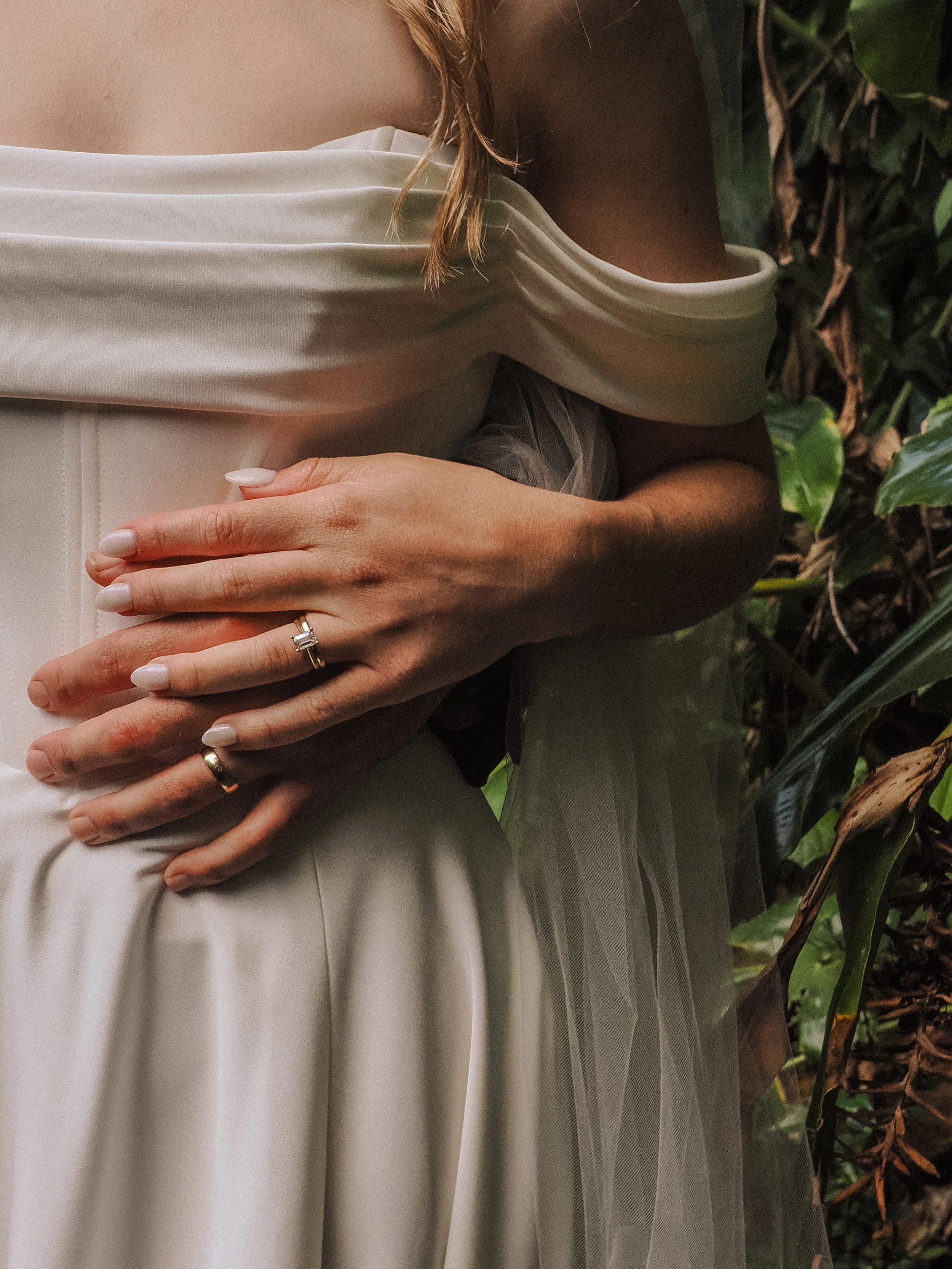 A woman in a white off-shoulder dress standing among green plants, with her hands crossed over her waist, wearing rings on her fingers.  Wedding Photography Waikato New Zealand. Wedding Photography Hamilton New Zealand. Northland Wedding Photography