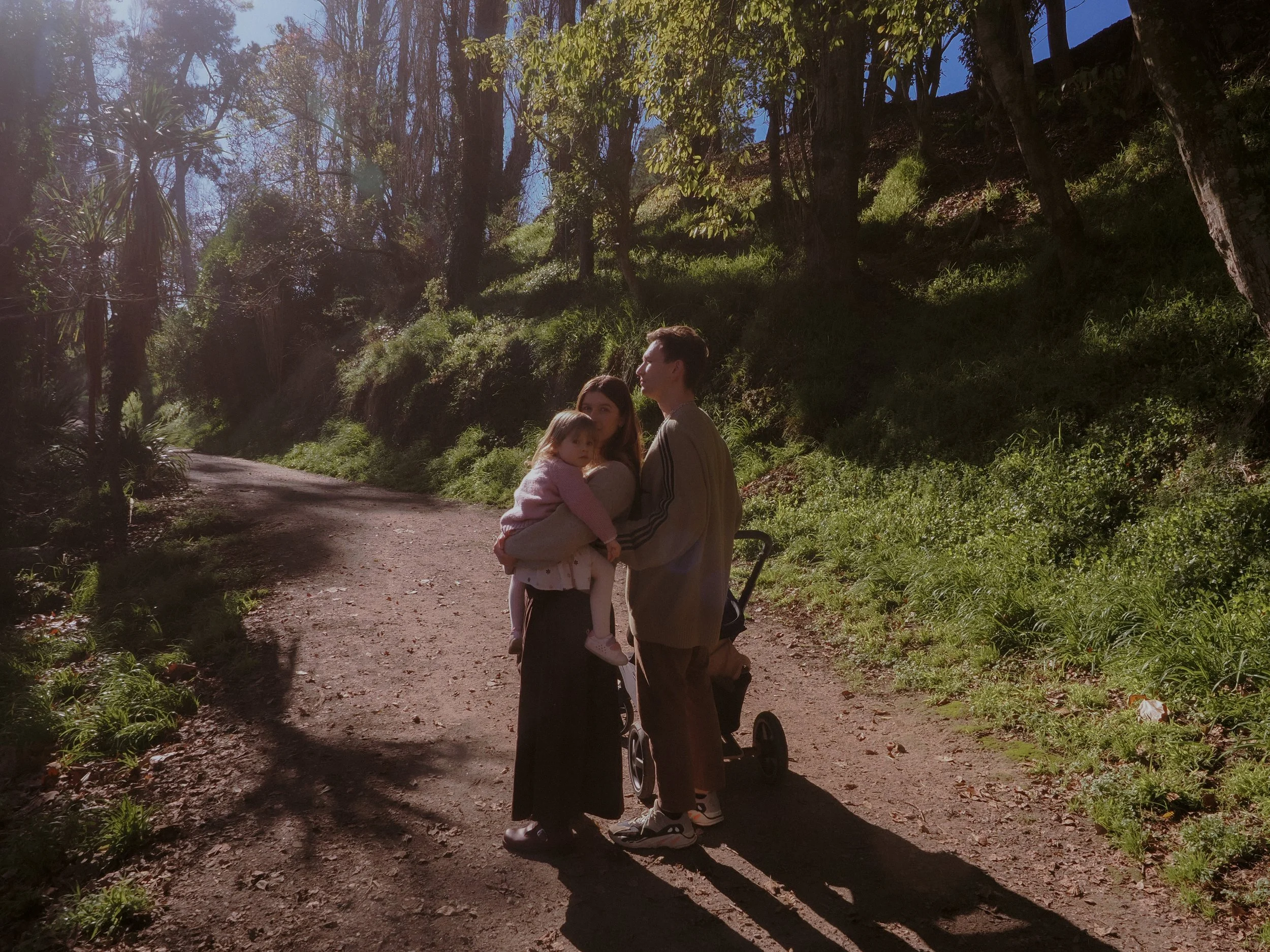A woman holding a young girl in a pink sweater, standing next to a man with a stroller, on a dirt path in a forested area during daytime.
