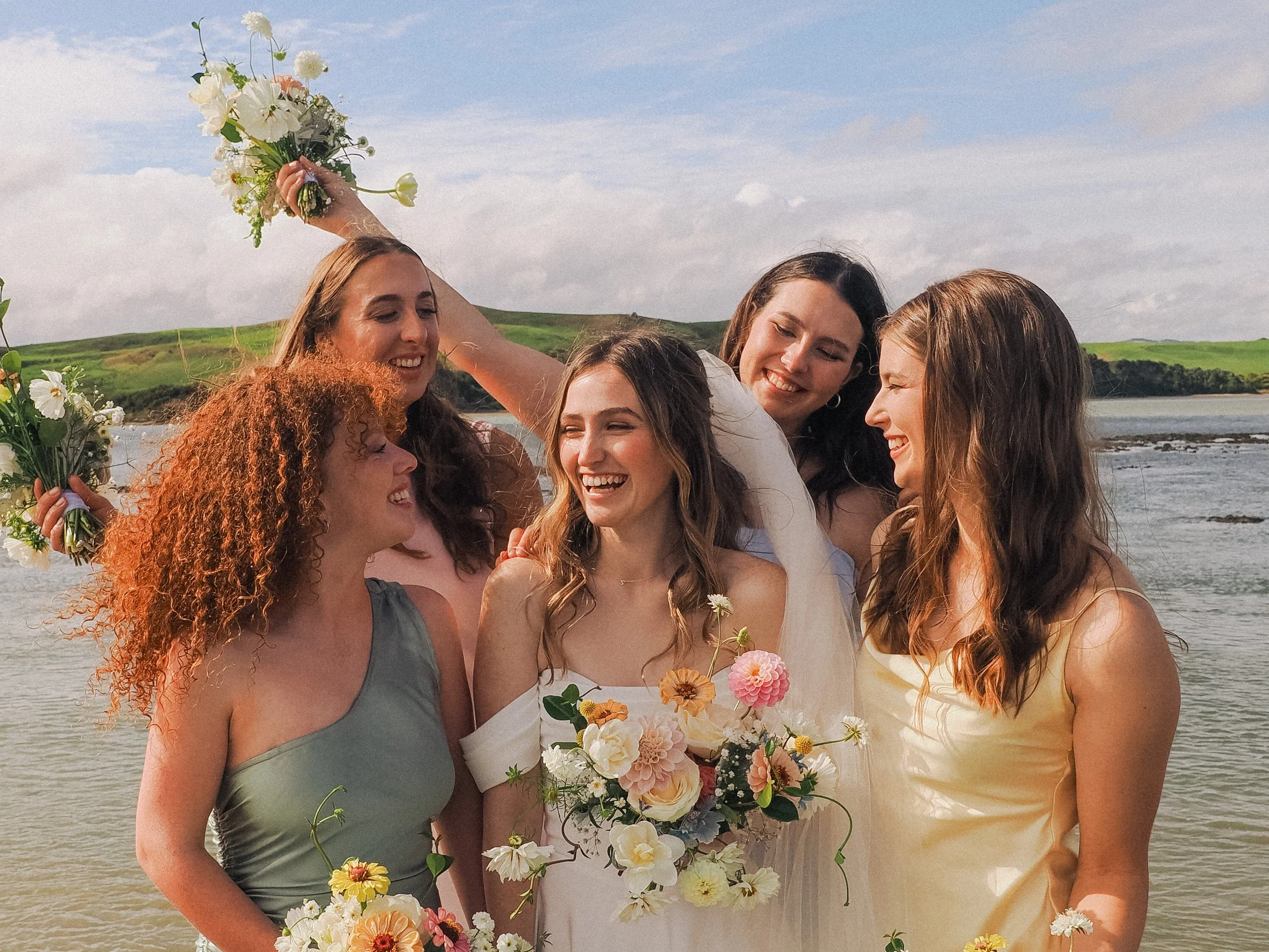 A group of five women on a beach, celebrating a wedding, with one holding a bouquet and another raising her arm in joy.  Wedding Photography Waikato New Zealand. Wedding Photography Hamilton New Zealand. Northland Wedding Photography