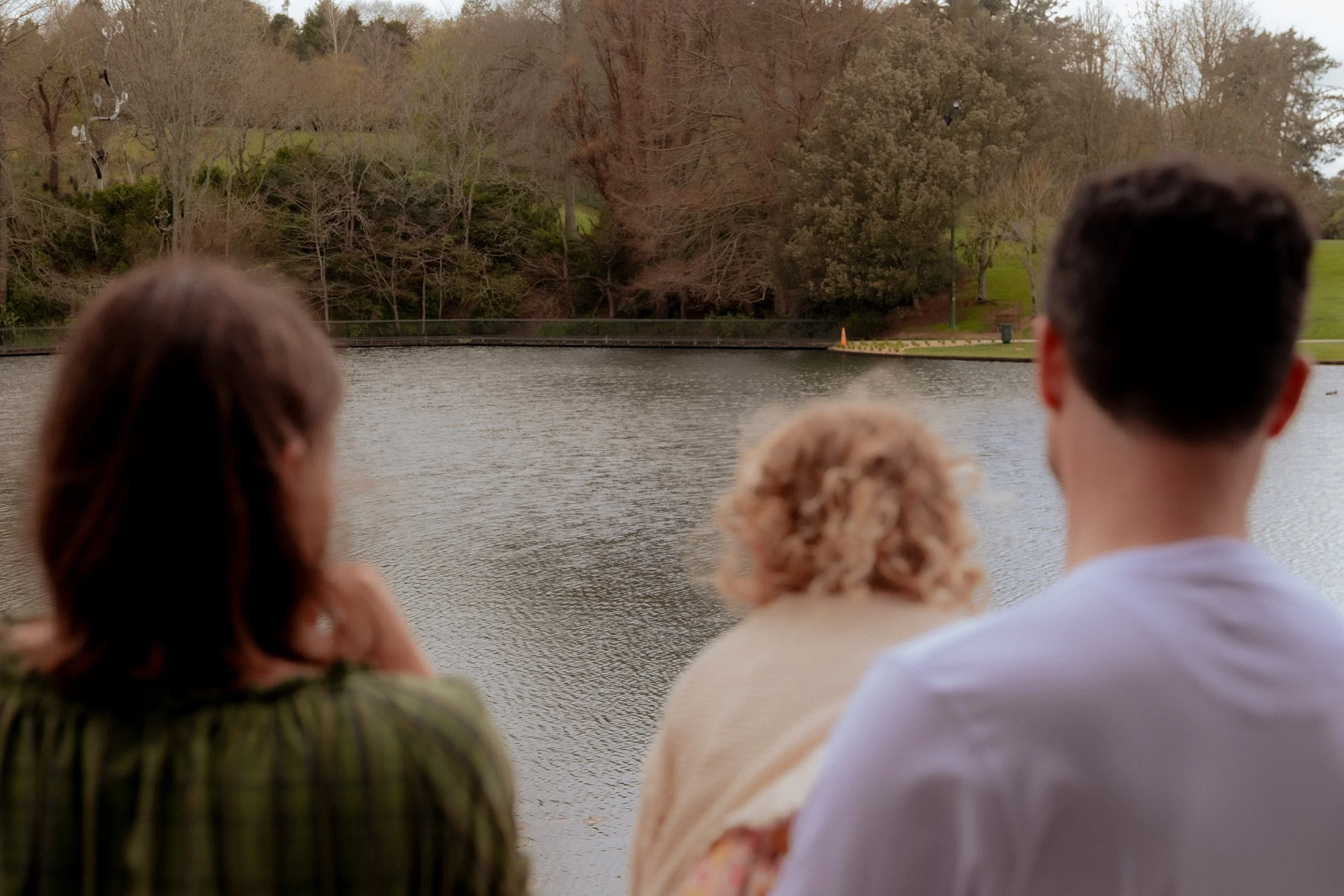 Three people sitting by the water, looking at a lake with trees in the background. Family/Maternity Photography Hamilton New Zealand