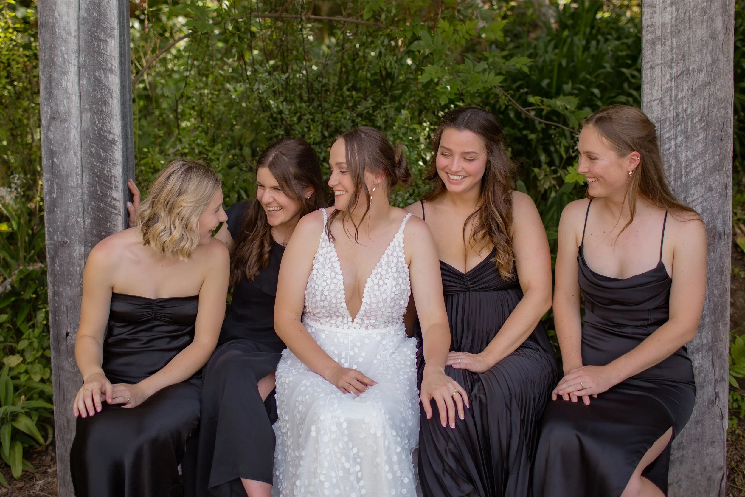 A group of five women, one in a white dress and four in black dresses, sitting outdoors on a wooden structure, smiling and laughing together.  Wedding Photography Waikato New Zealand. Wedding Photography Hamilton New Zealand. Northland Wedding Photo