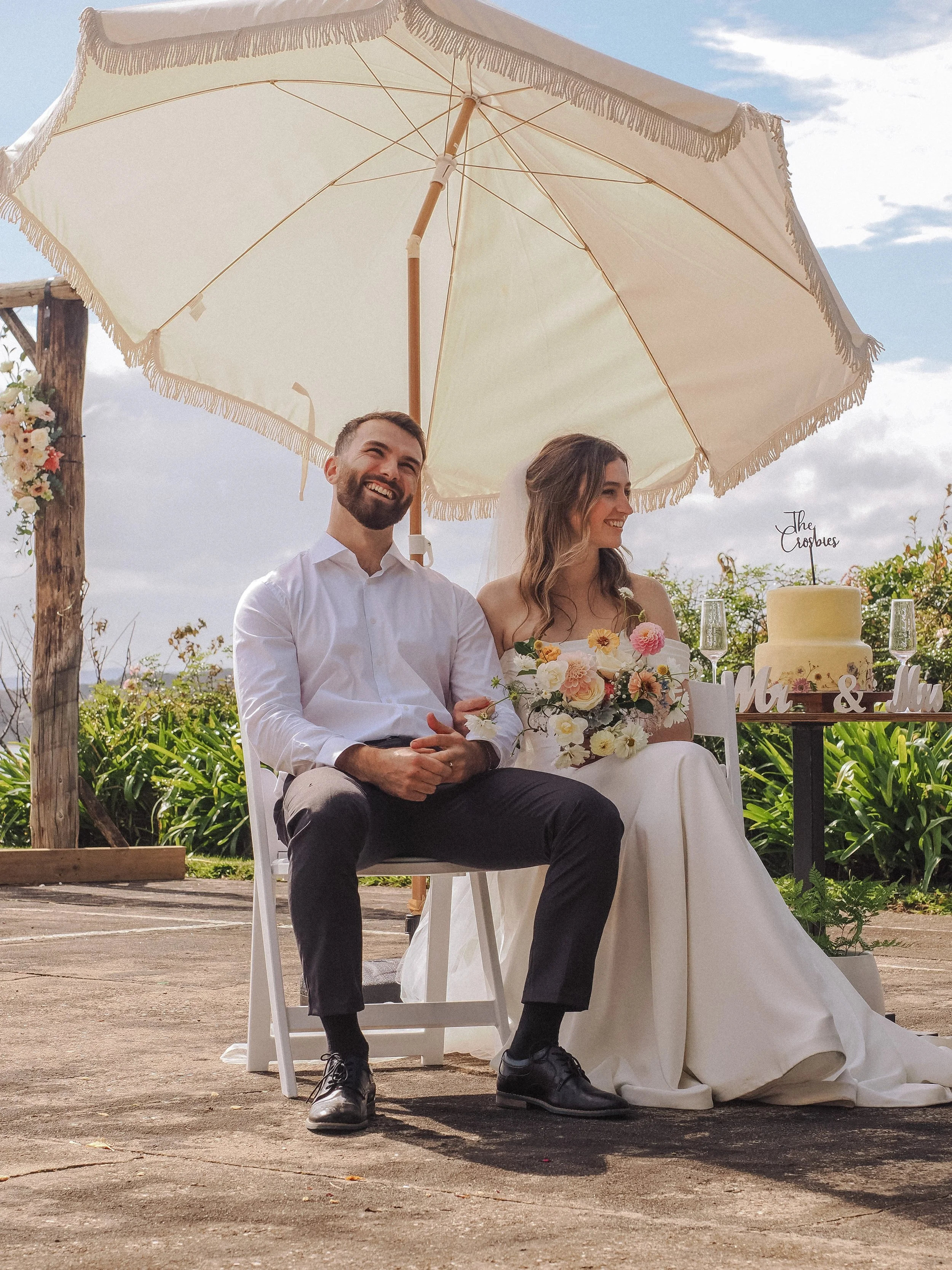 A bride and groom sitting under a large white umbrella during their outdoor wedding ceremony, with a wedding cake and floral decorations visible behind them.  Wedding Photography Waikato New Zealand. Wedding Photography Hamilton New Zealand. 