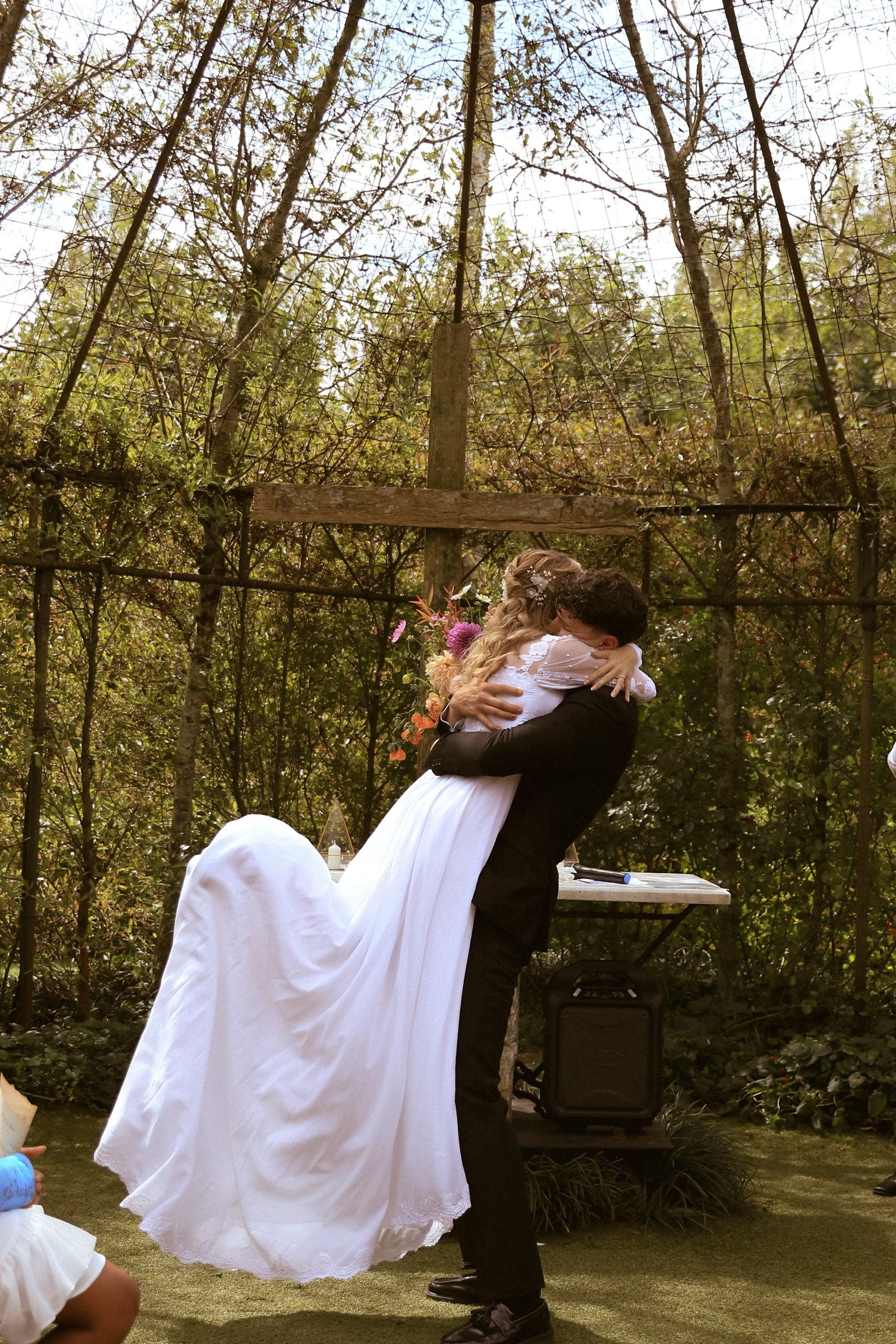 A bride and groom kissing outdoors at their wedding ceremony, surrounded by trees, with the groom lifting the bride off the ground.