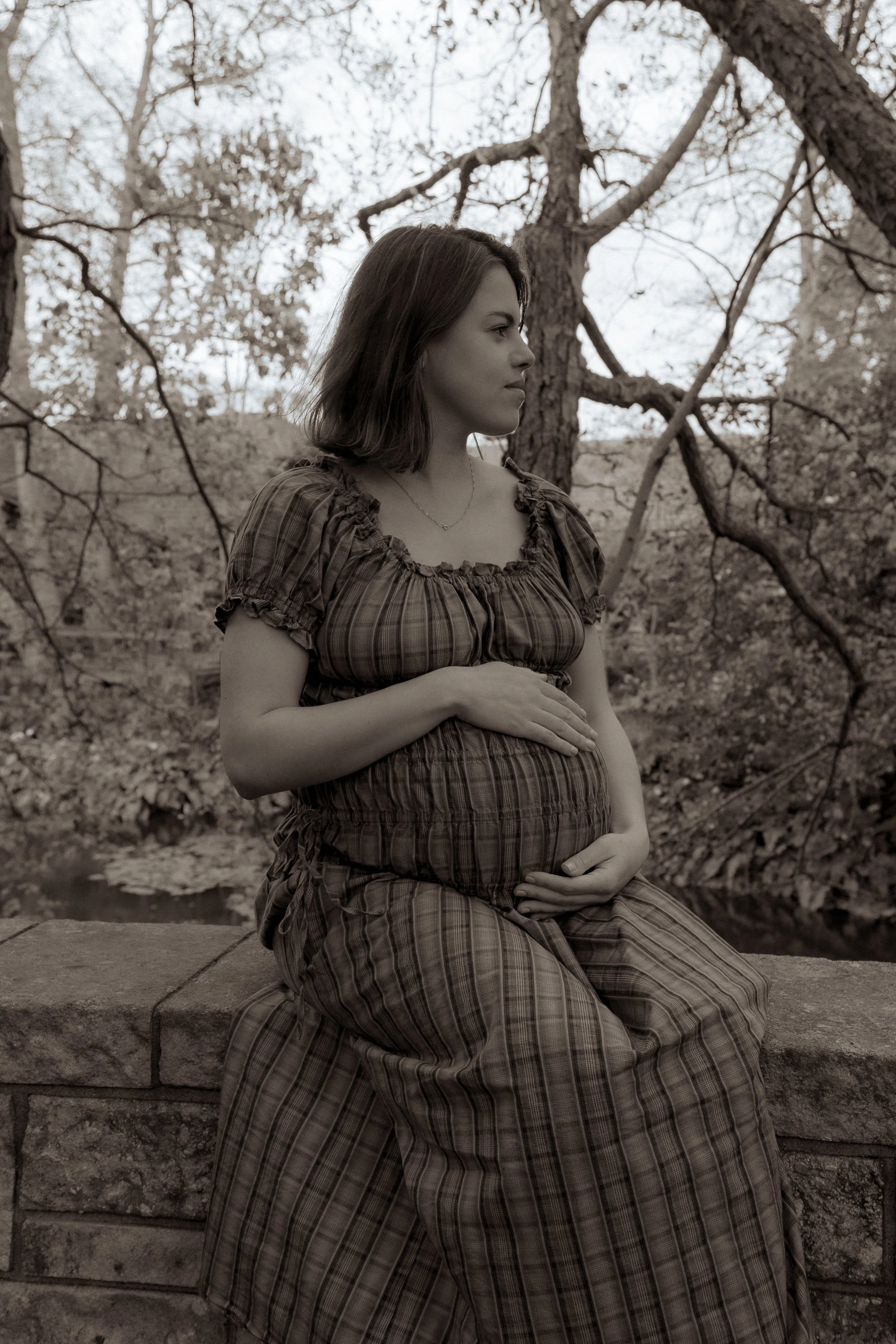 A pregnant woman sitting outdoors on a stone ledge, gently holding her belly, with trees and a small stream in the background. Maternity Photography Hamilton New Zealand