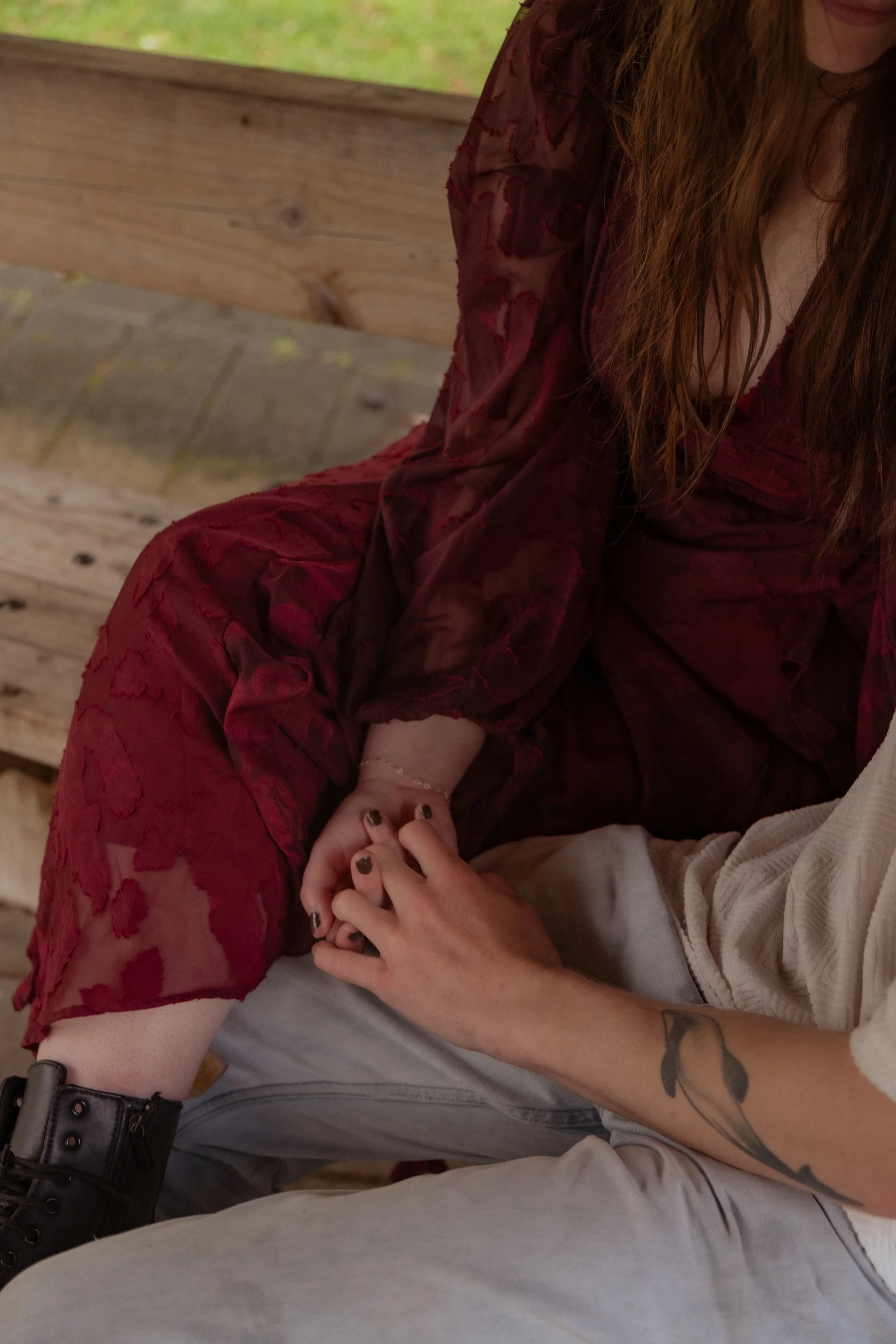 A person with long red hair wearing a sheer, dark red dress sitting on a wooden bench, holding hands with another person. Couples photography Raglan New Zealand. Engagement photography Hamilton New Zealand. Whangarei New Zealand Couples photographer