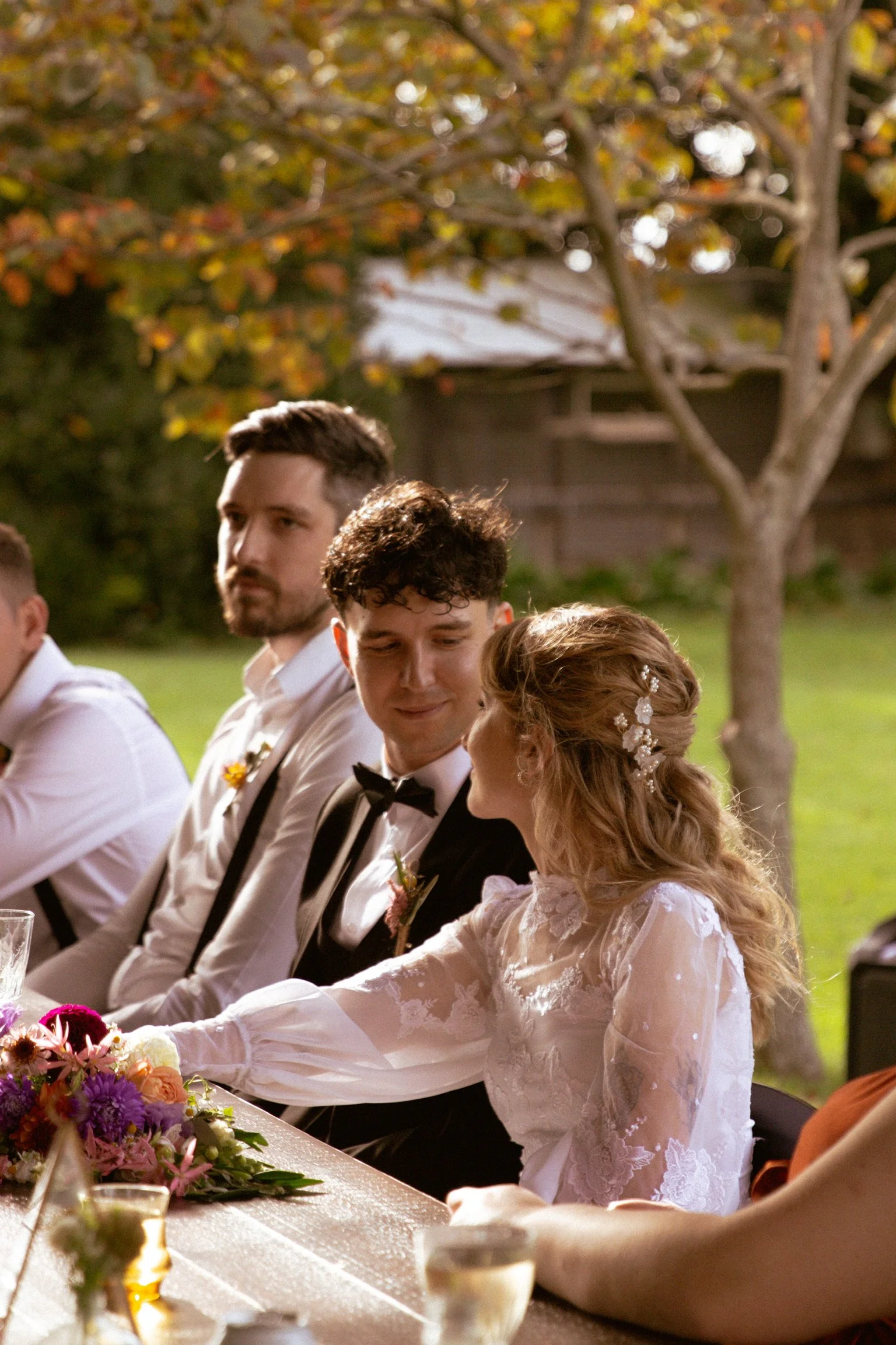 A couple at their wedding reception sharing a tender moment, surrounded by friends, with a tree in the background during sunset or evening. Wedding Photography Waikato New Zealand. Wedding Photography Hamilton New Zealand