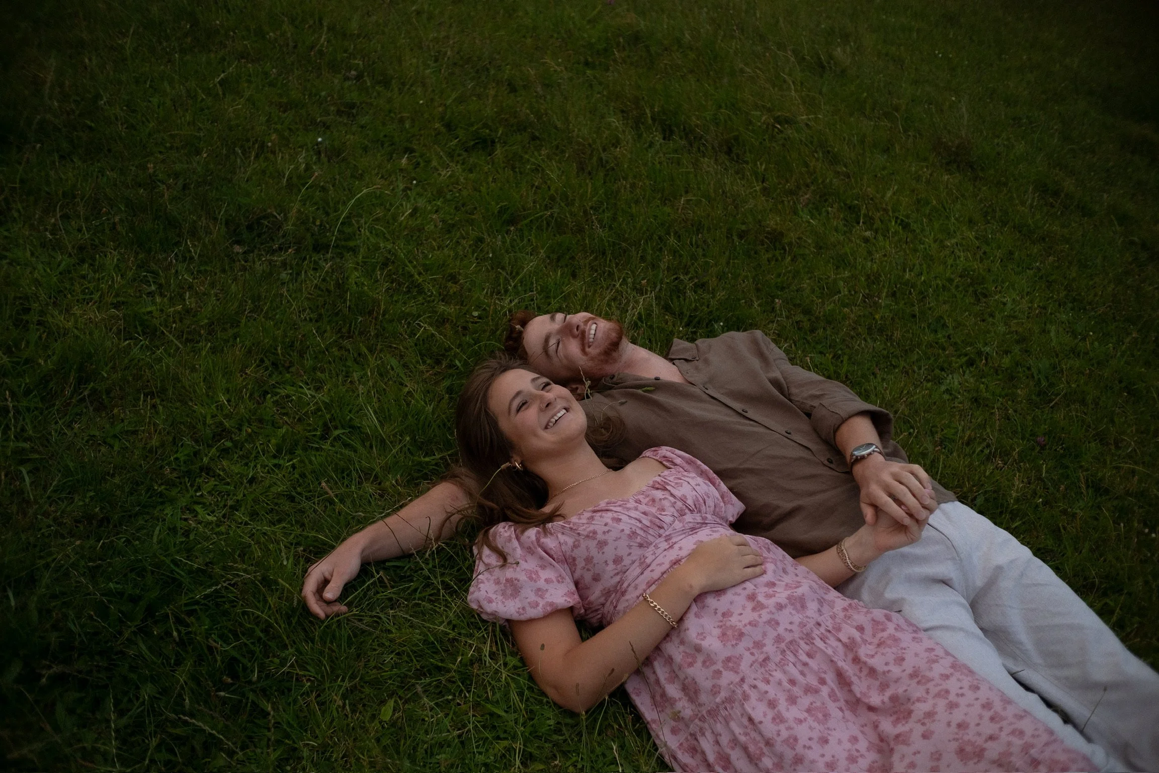 A young woman and a man lying on the grass, smiling and holding hands, enjoying a moment together. Couples photography Raglan New Zealand. Engagement photography Hamilton New Zealand. Whangarei New Zealand Couples photographer
